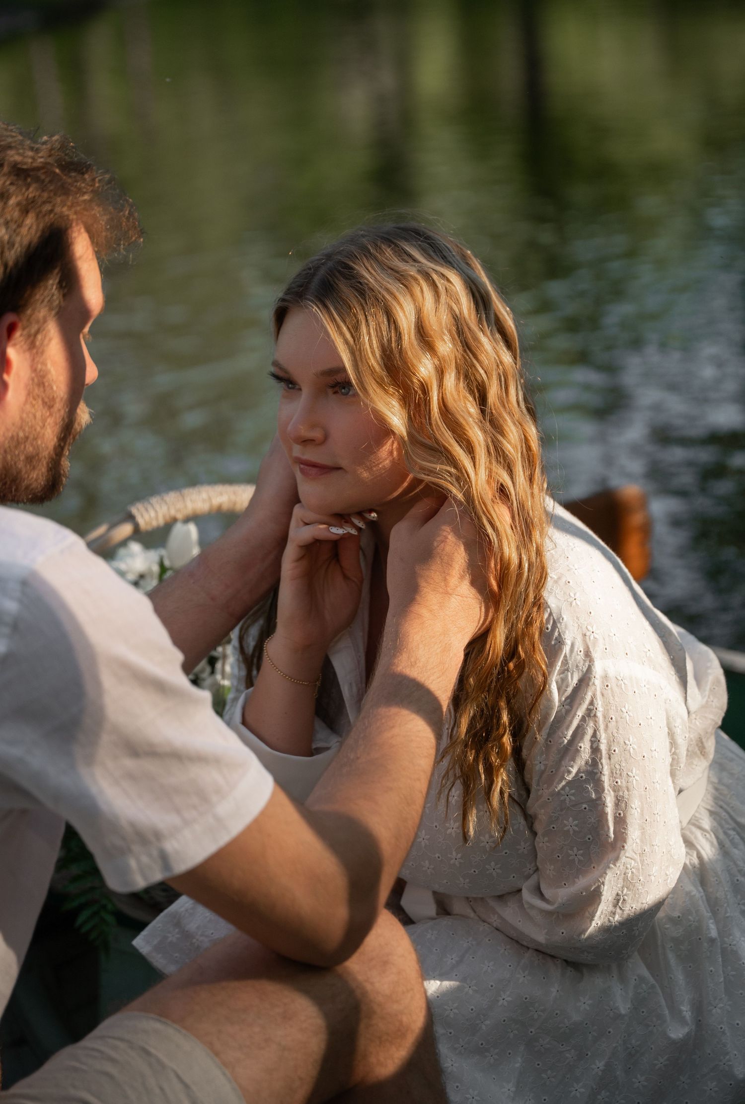 A man tenderly holds a woman's face near a serene lake, both dressed in light linen clothing.