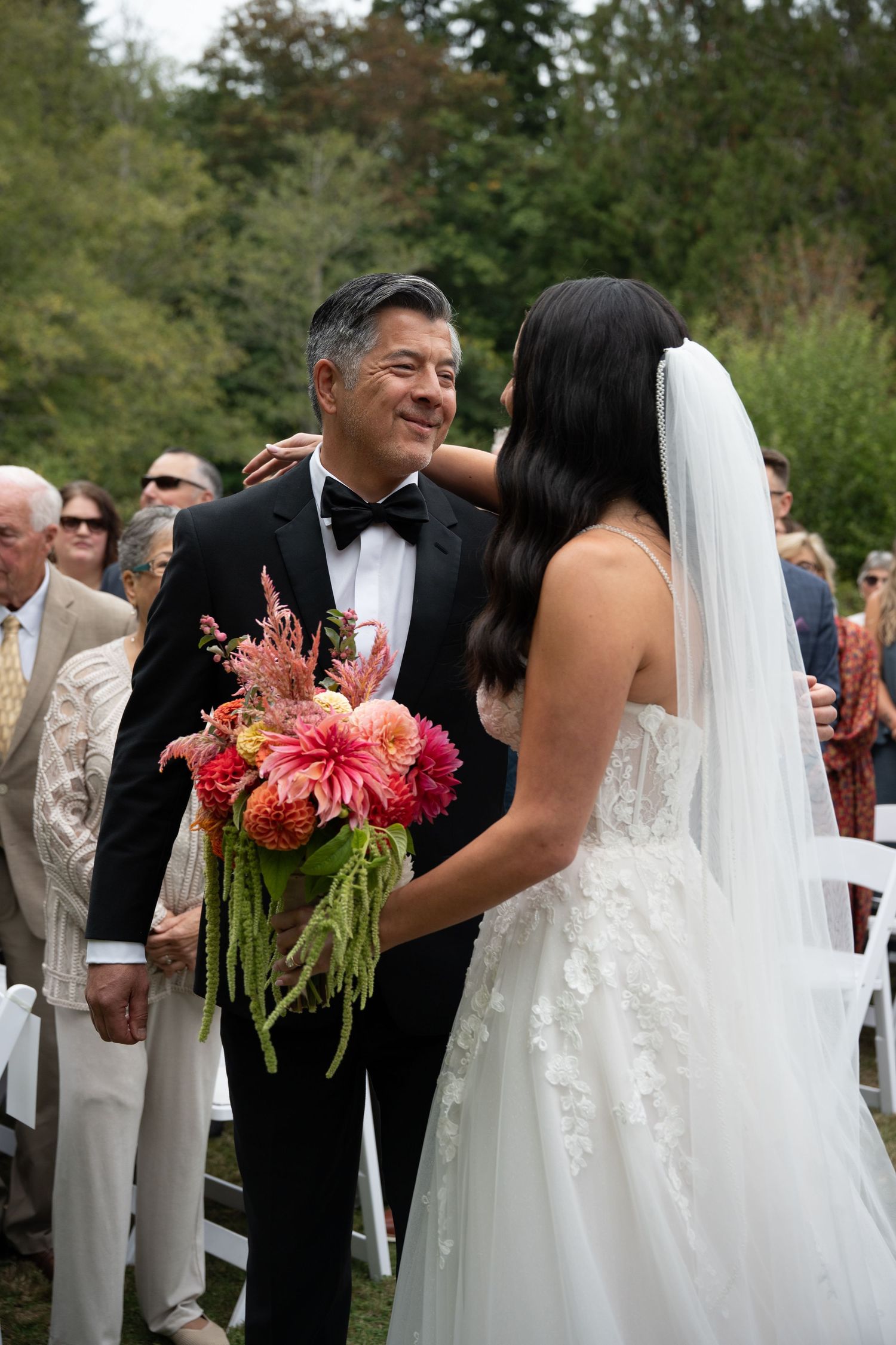 Bride in white lace gown and veil shares a tender moment with man in tuxedo holding pink dahlia bouquet at outdoor wedding.