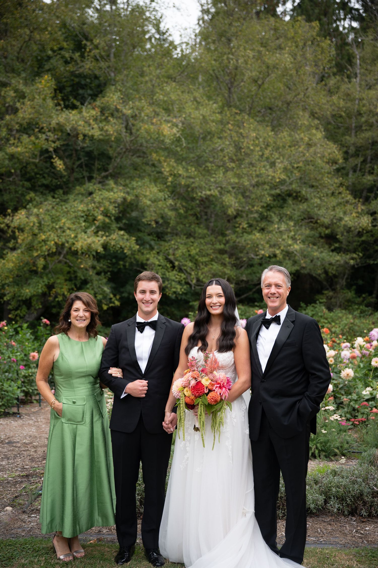 Bride and groom pose with family in garden setting, wearing formal attire with floral bouquet.