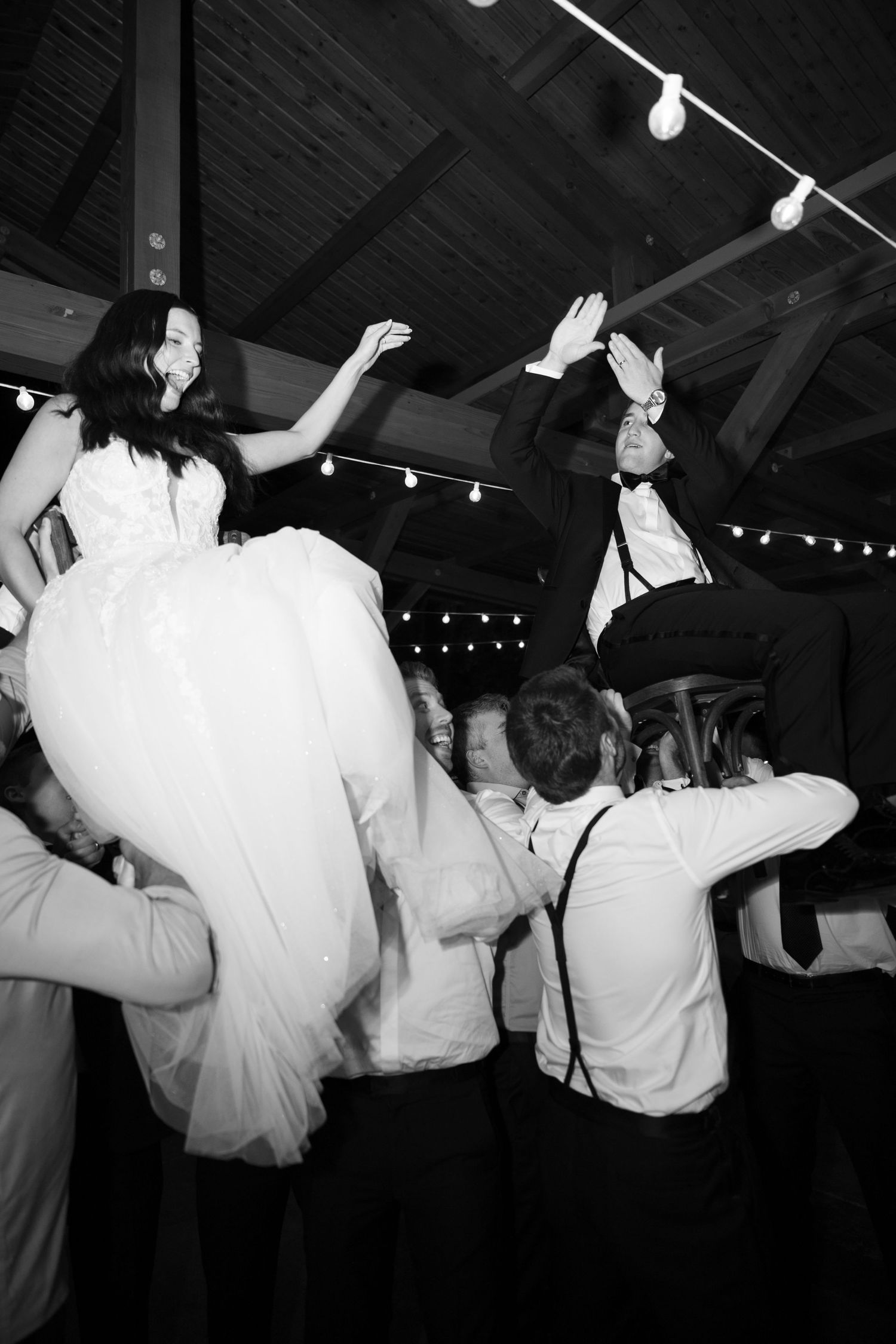 Black and white photo of bride and groom being lifted by guests during lively wedding reception dancing.