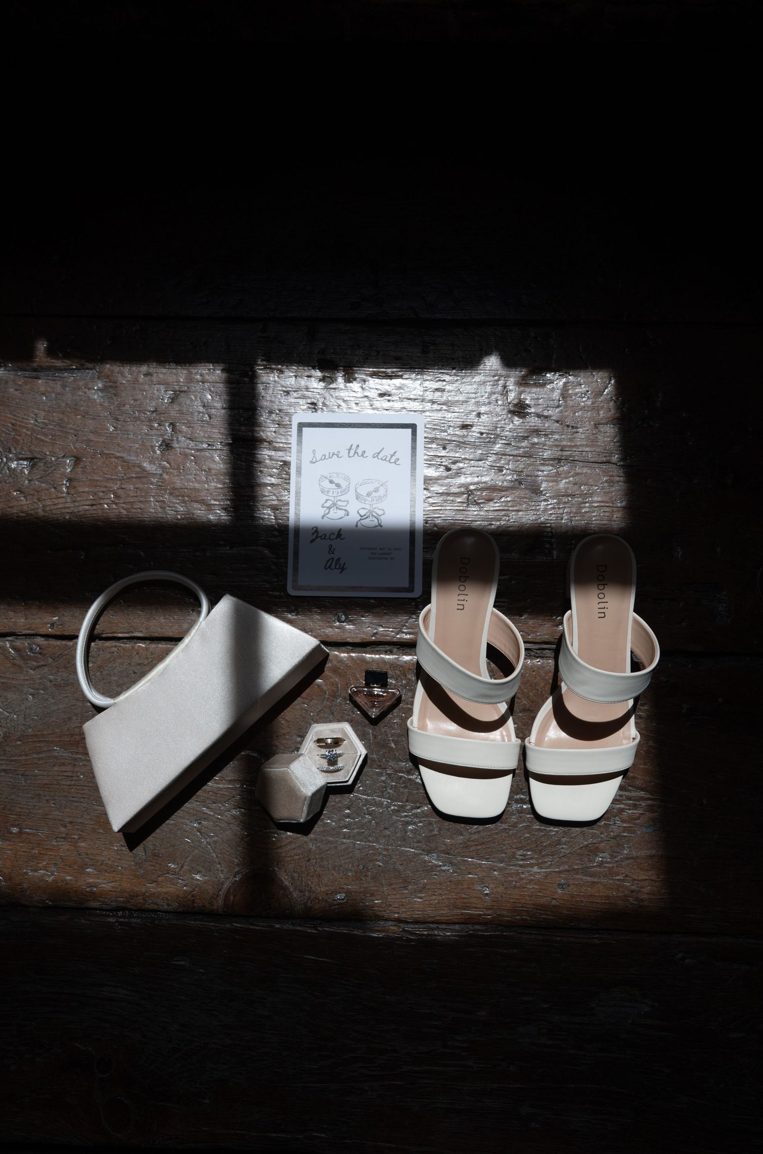 Wedding accessories flatlay featuring white heels, clutch, ring, and save the date card in dramatic lighting.