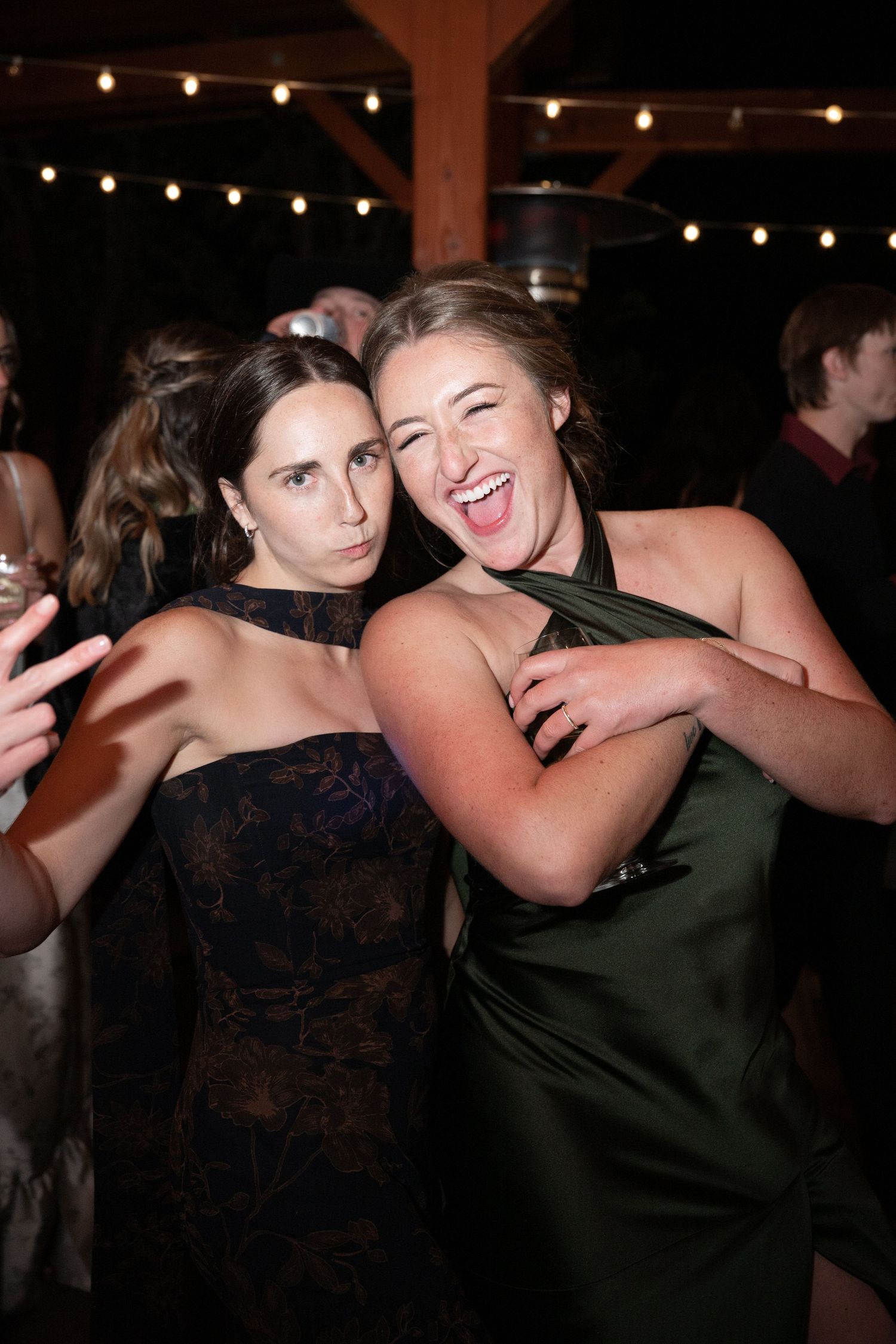 Two women in formal dresses laugh and embrace on the dance floor at wedding reception.