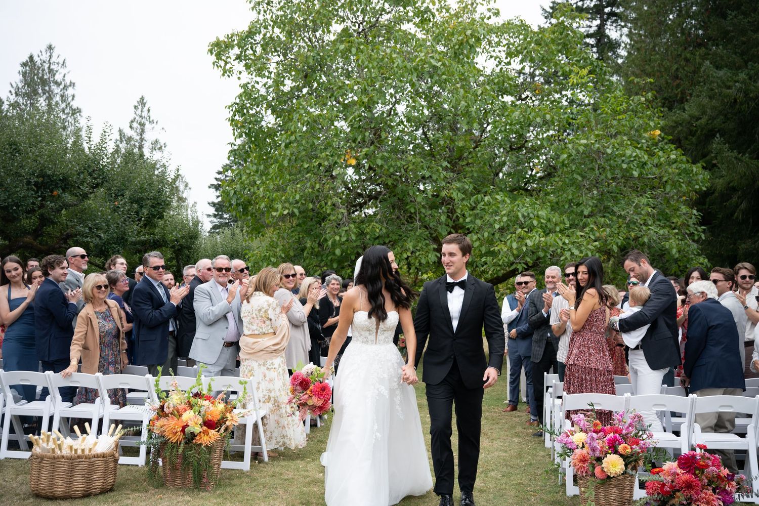 Bride and groom walk down outdoor wedding aisle surrounded by guests and colorful floral arrangements.
