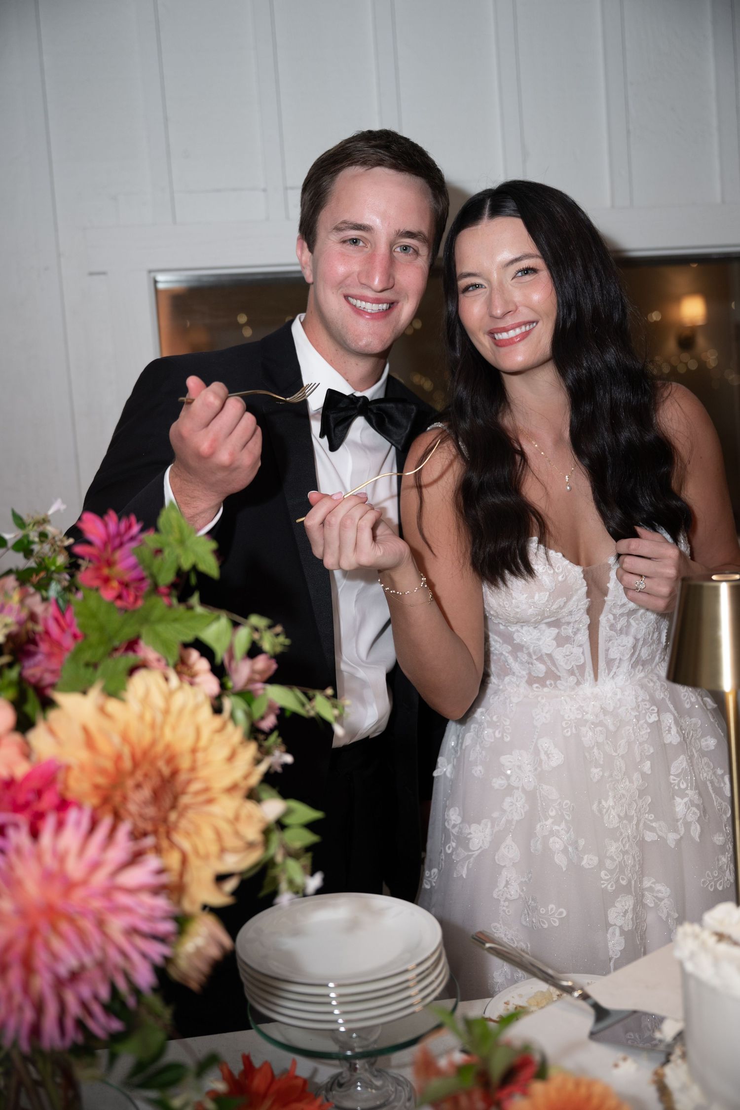 Smiling bride and groom in formal attire cut their wedding cake surrounded by colorful dahlia flowers.
