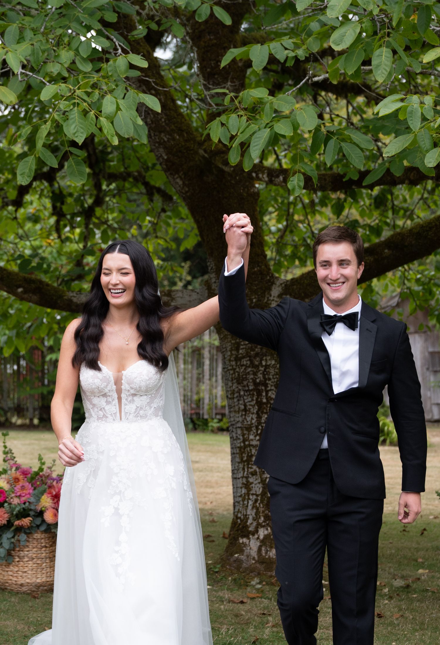 Bride and groom raising joined hands under a large tree, wearing wedding gown and black tuxedo.