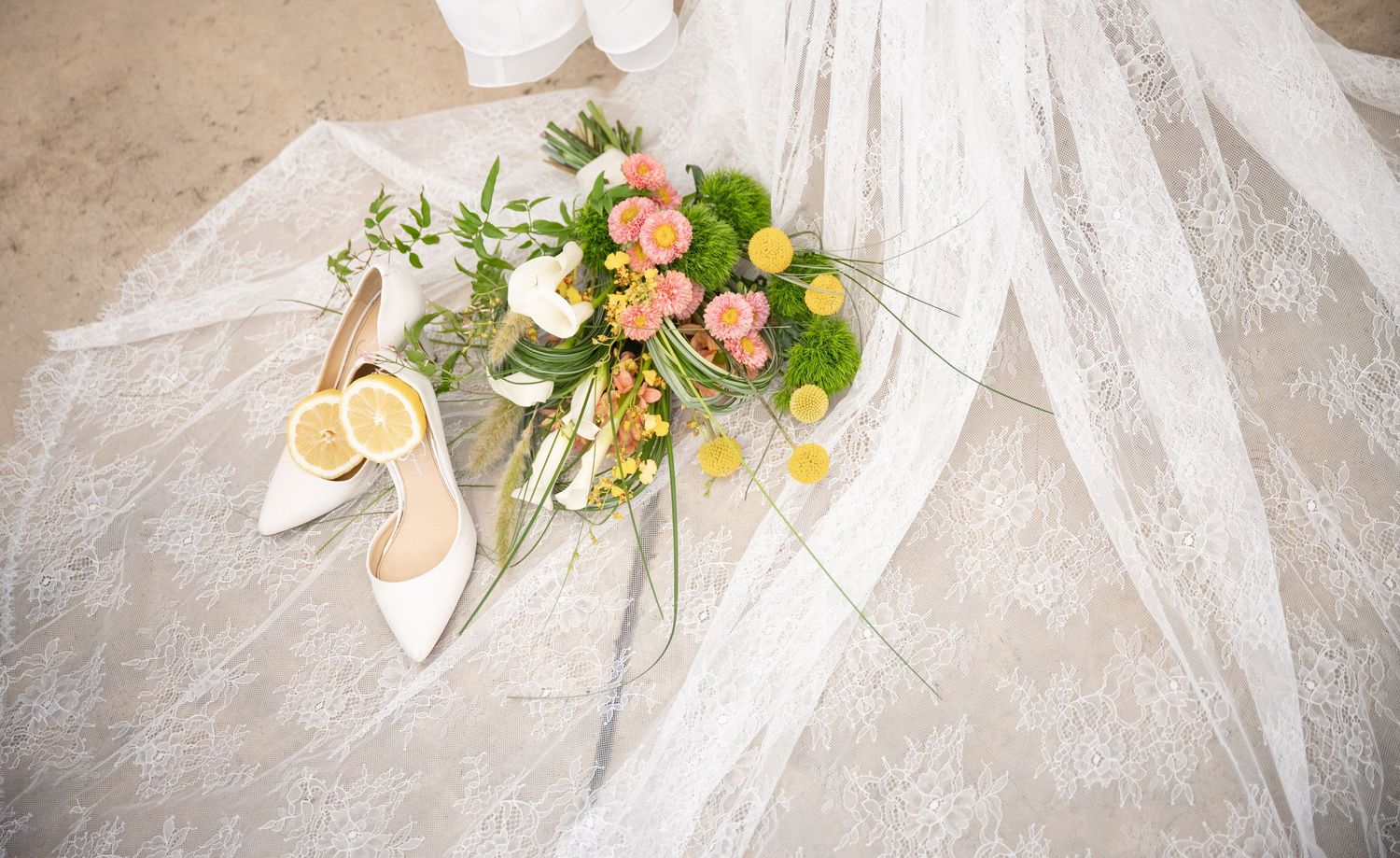 White bridal heels with lemon slices beside a lush pink and green wedding bouquet on a delicate lace veil.