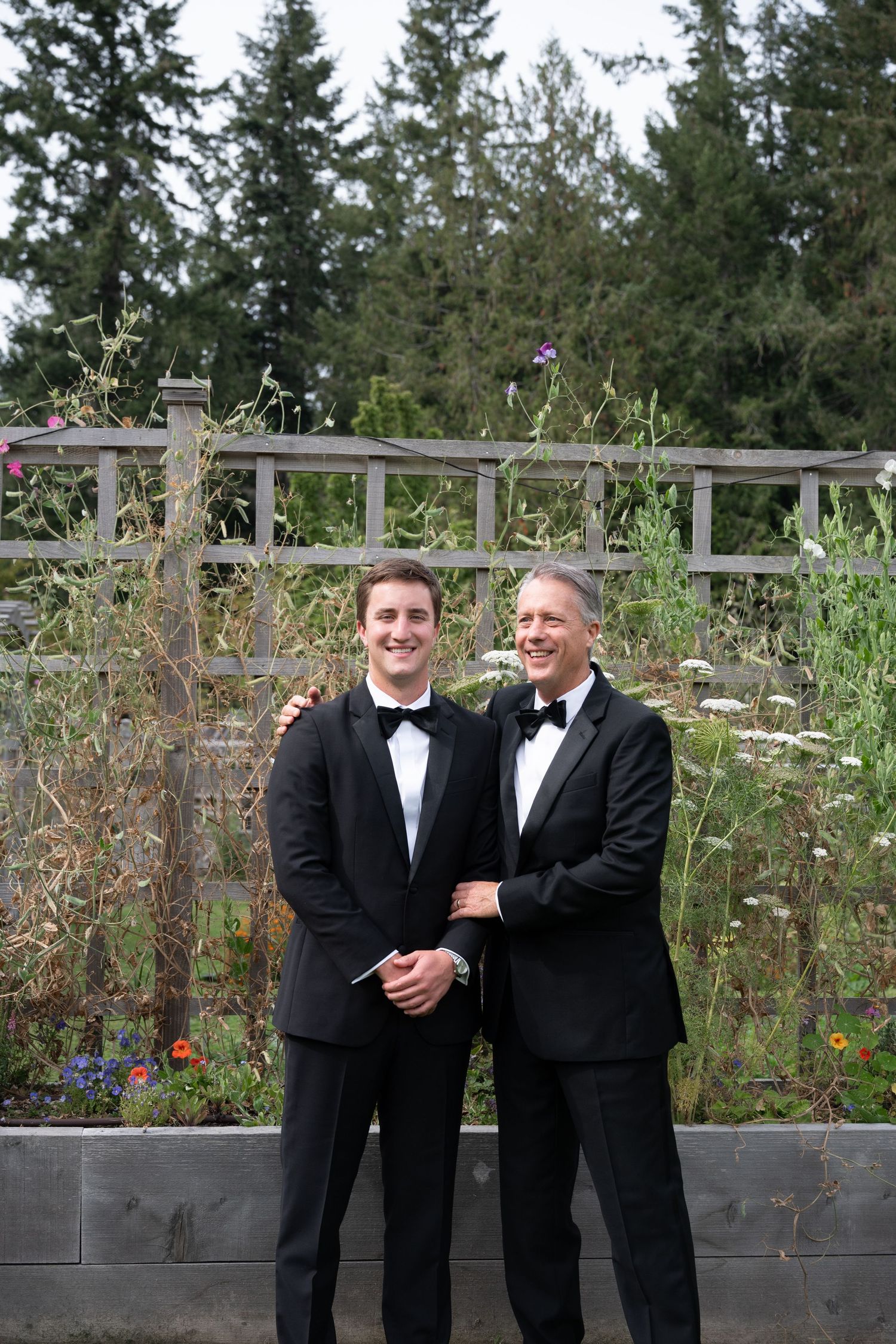 Two men in black tuxedos smile together outdoors at a wedding, posing in front of a wooden garden fence.