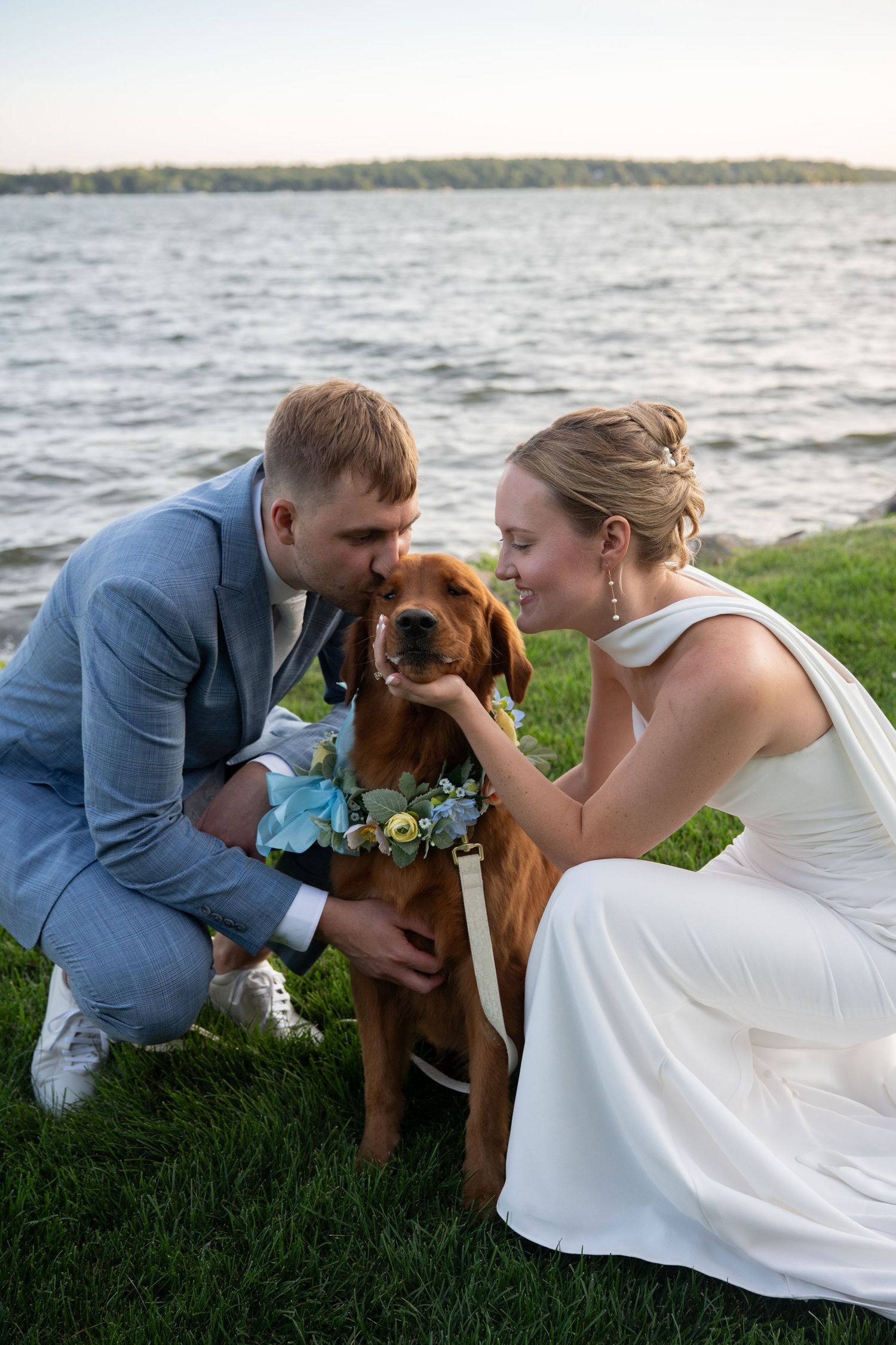 Bride and groom crouch together to pet their golden retriever wearing a floral collar by the lake.