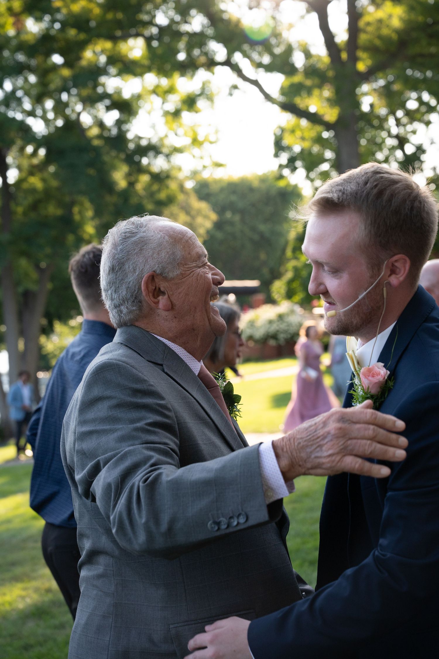 Elderly man in gray suit shares joyful moment with young groom wearing boutonniere at outdoor wedding.