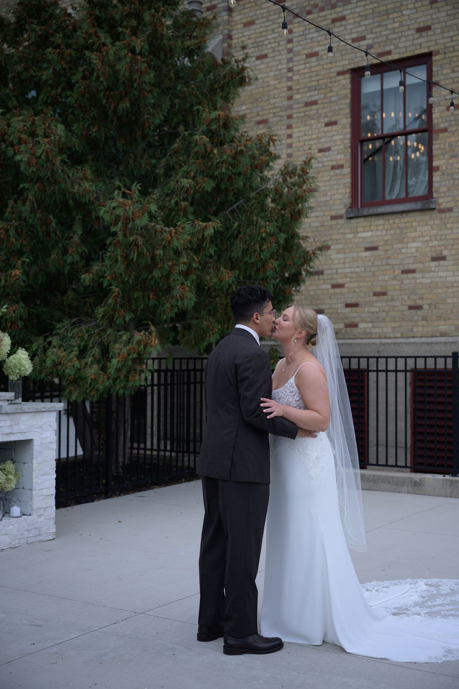 Bride and groom share a kiss outdoors near a brick building, bride wearing white gown with veil.