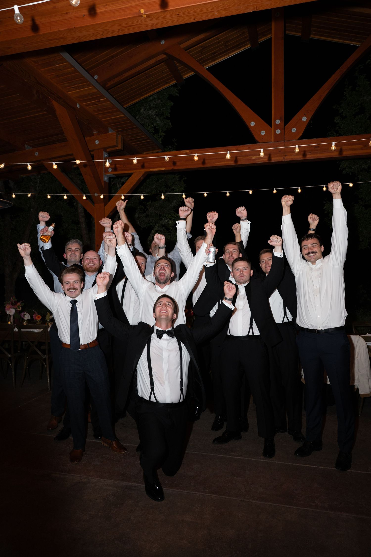 Groom and groomsmen cheer with raised fists under string lights at a wedding reception celebration.
