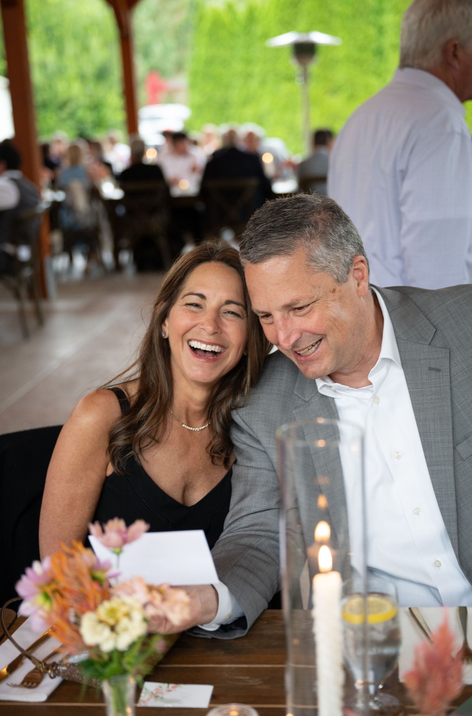 Couple laughing together at an outdoor wedding reception, seated at a decorated table with flowers and candles.