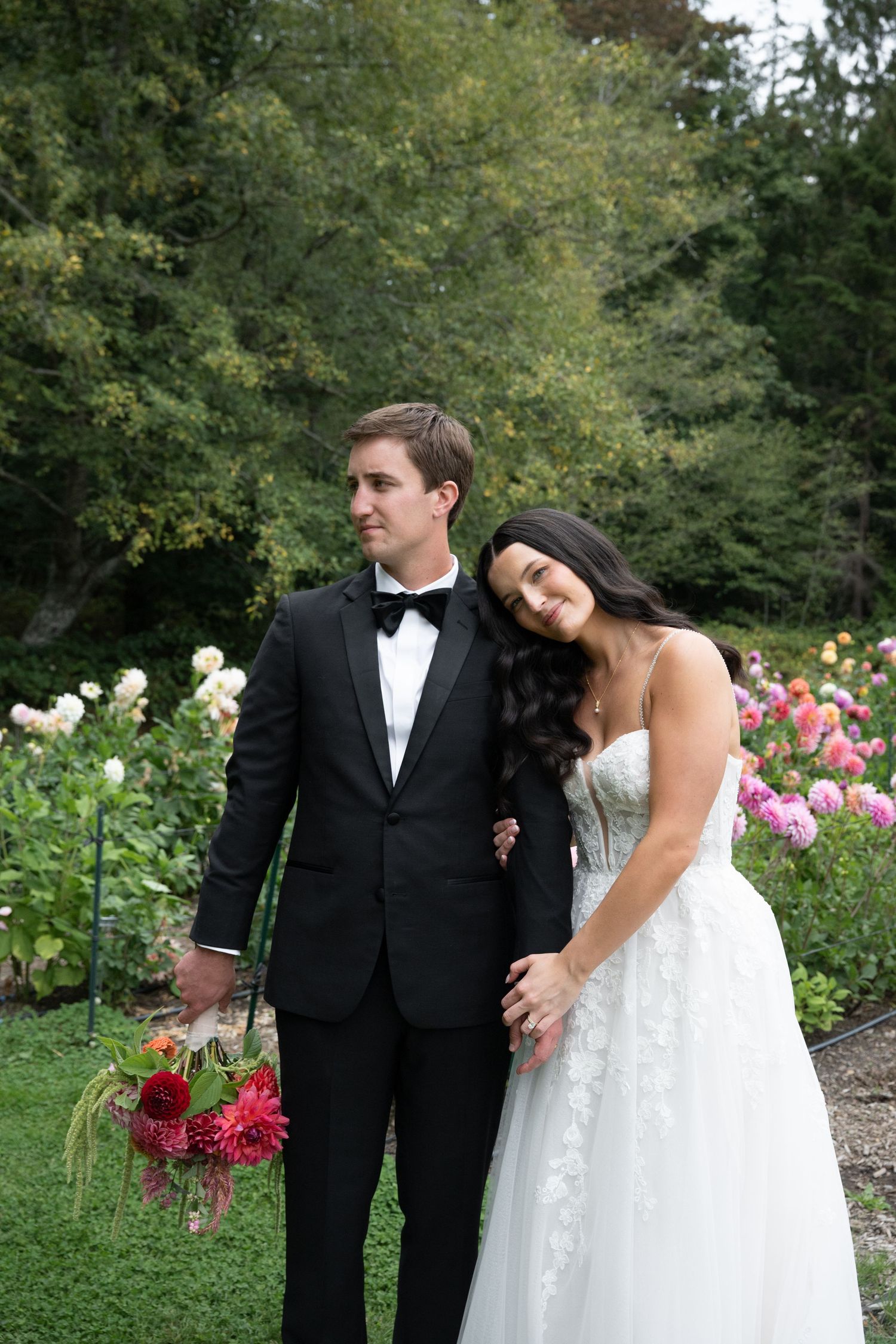 Bride in white lace gown leans on groom in black tuxedo holding red dahlia bouquet in garden.