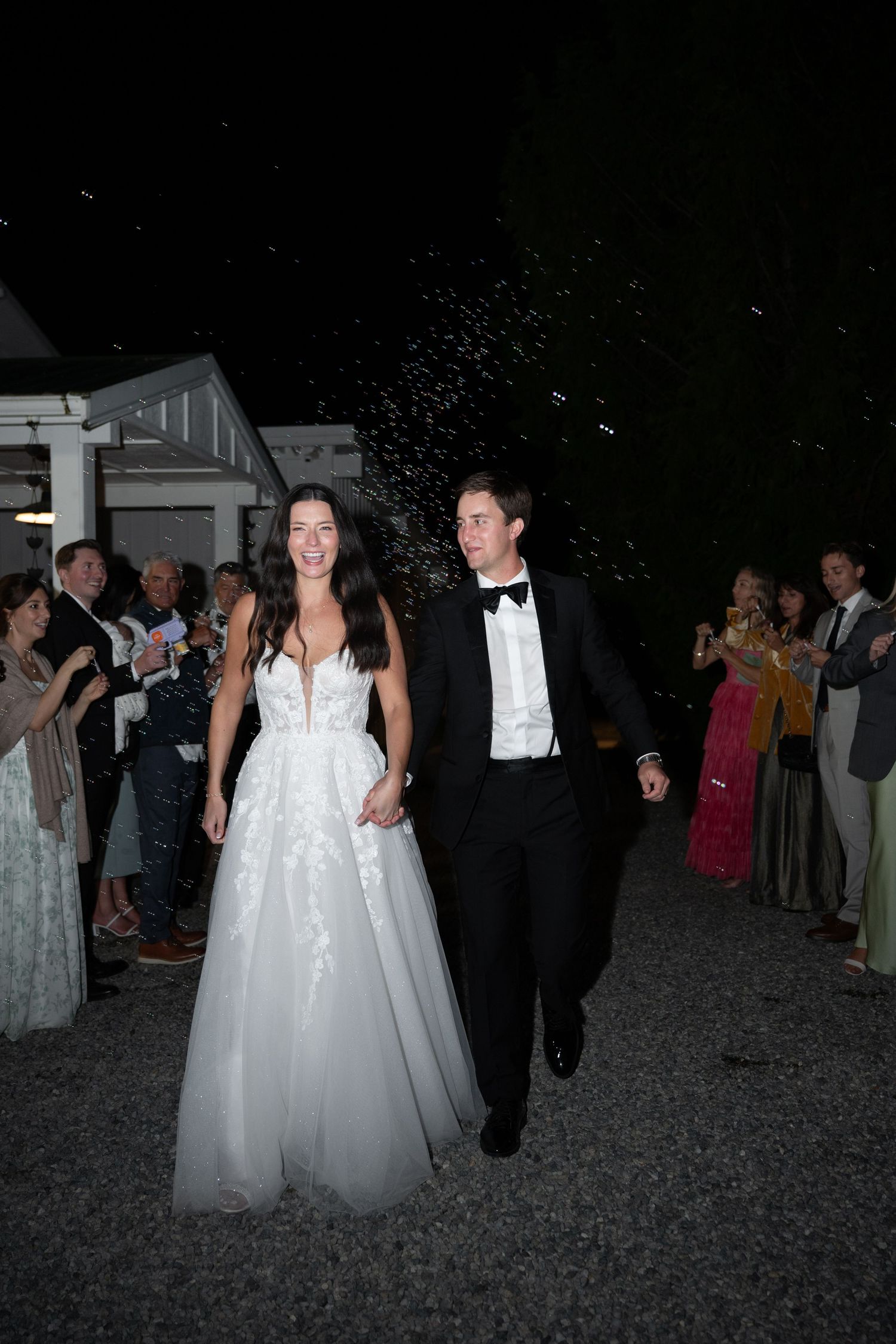 Bride and groom walking hand in hand through celebratory send-off with guests cheering at night wedding exit.