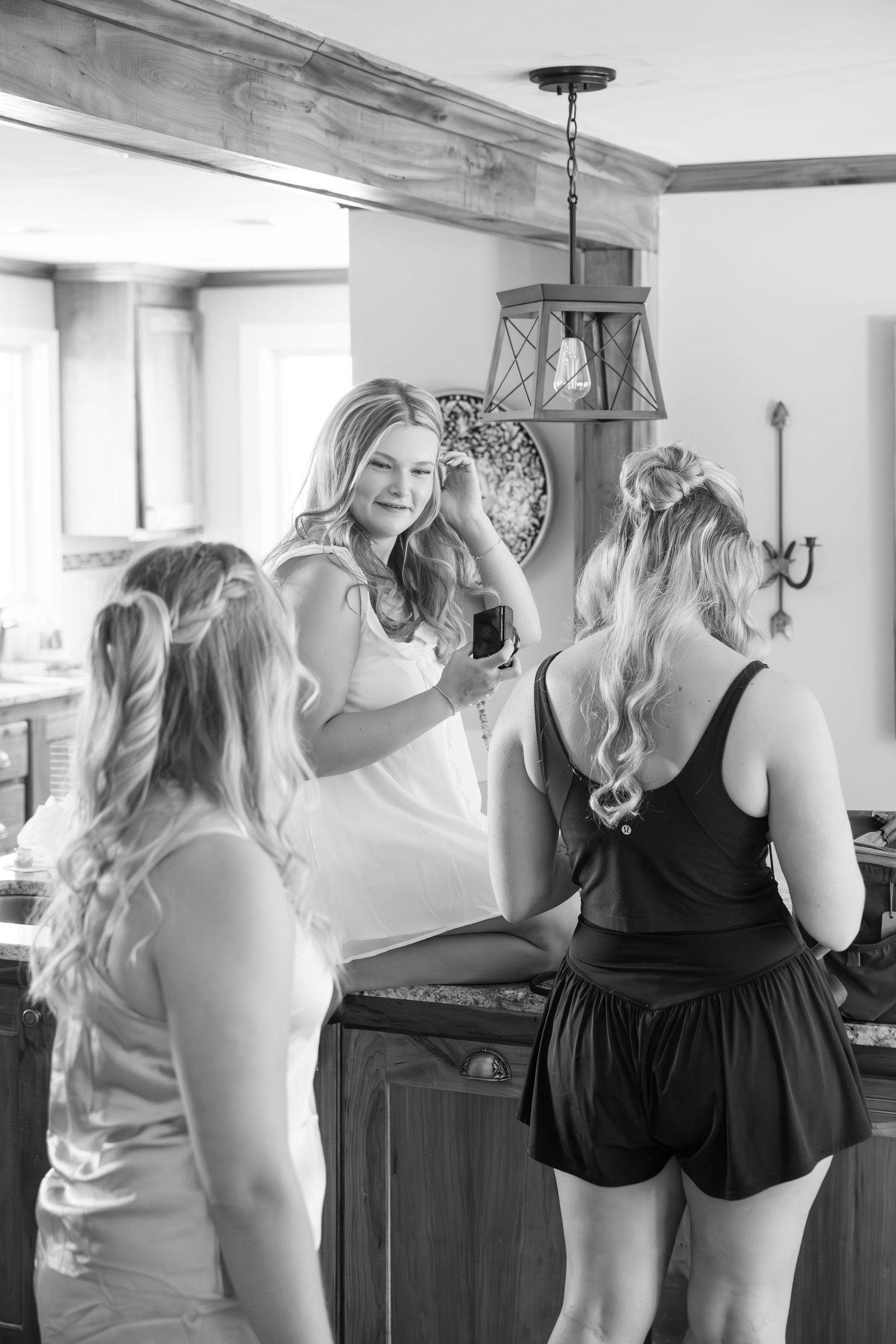 Black and white photo of three women with long blonde hair getting ready together in a rustic kitchen interior.