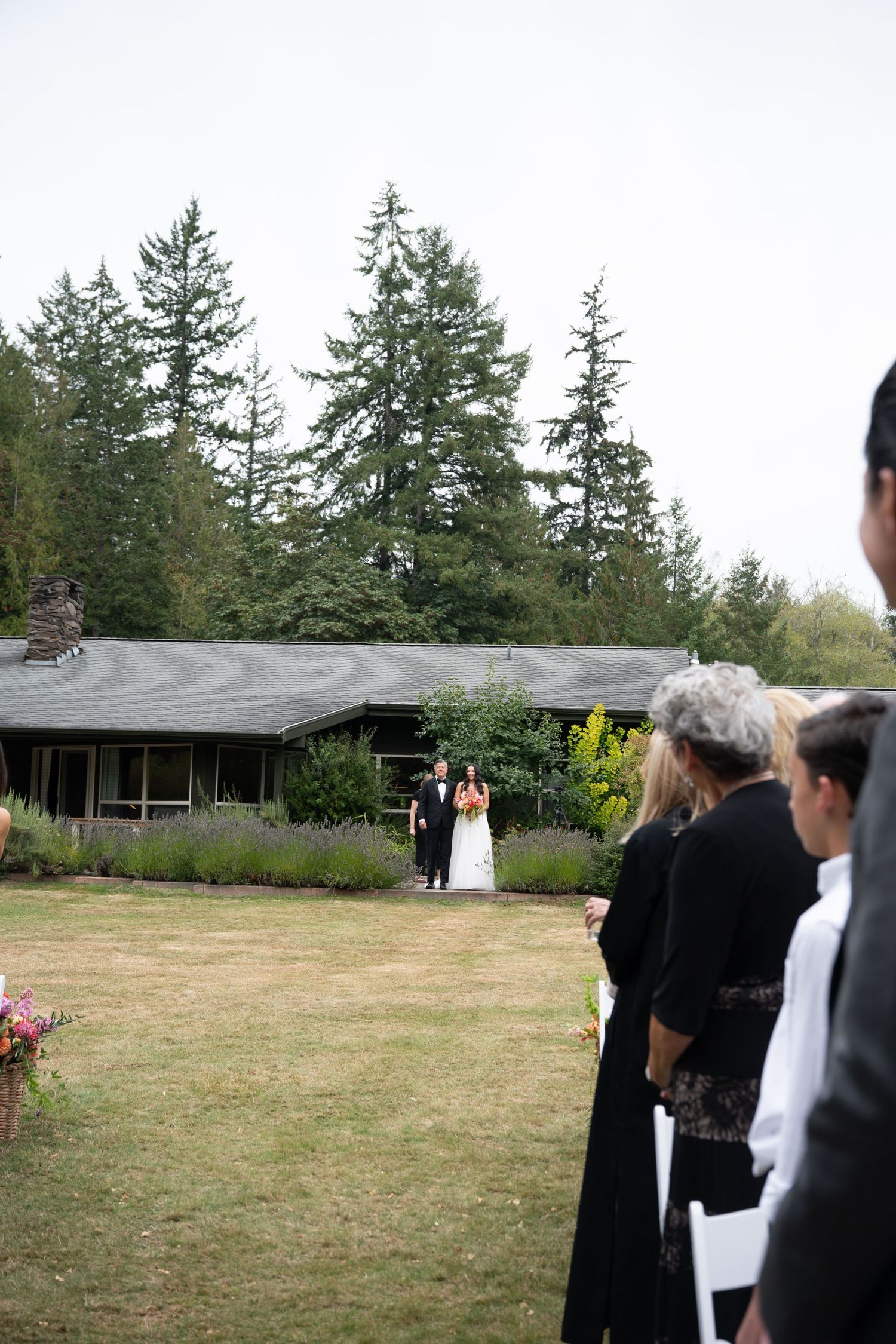 Bride and groom walking down outdoor aisle toward seated wedding guests surrounded by tall evergreen trees.