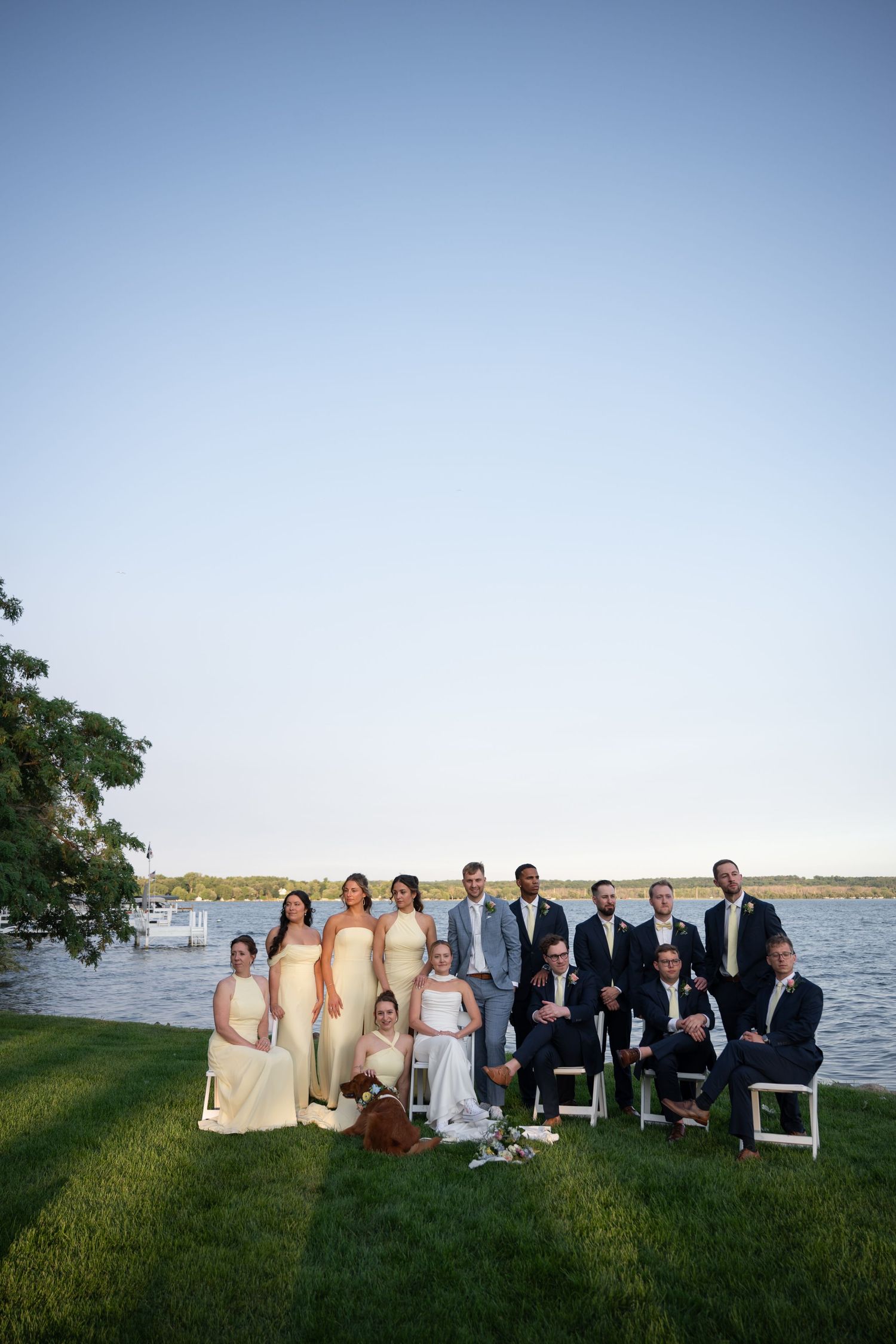 Wedding party poses together on green lawn by a lake at dusk, bridesmaids in yellow gowns.