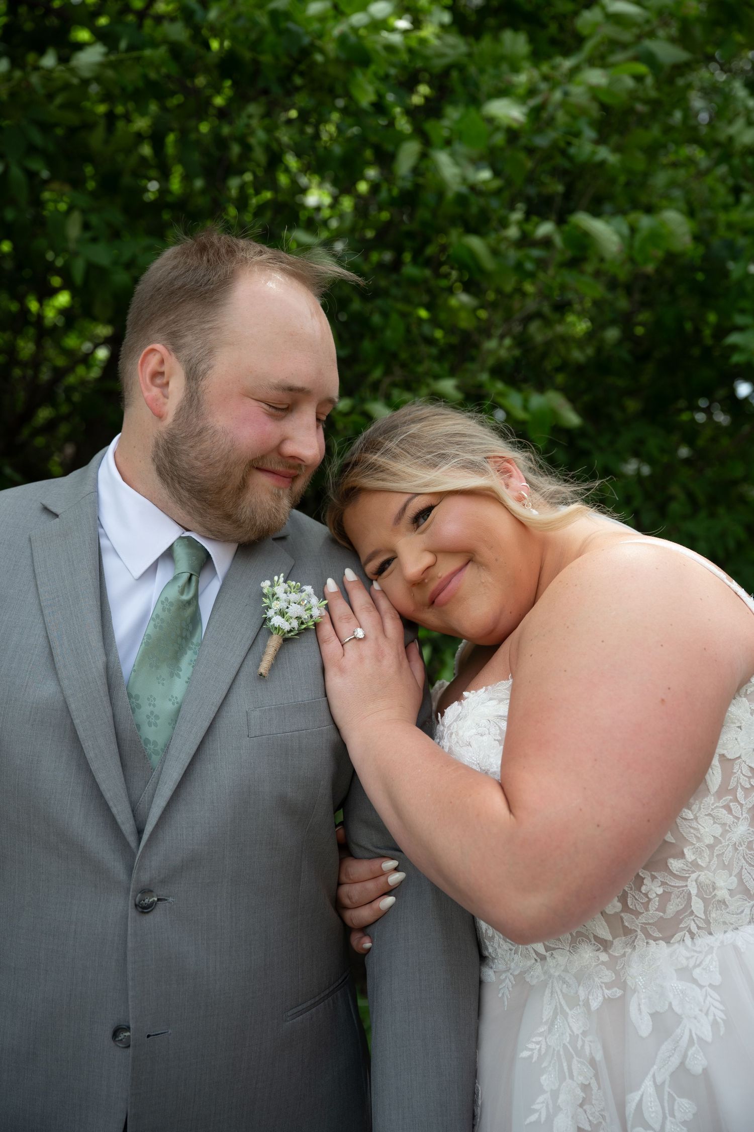 A bride and groom share a tender moment outdoors, she rests her head on his chest wearing a lace gown, he wears a gray suit.