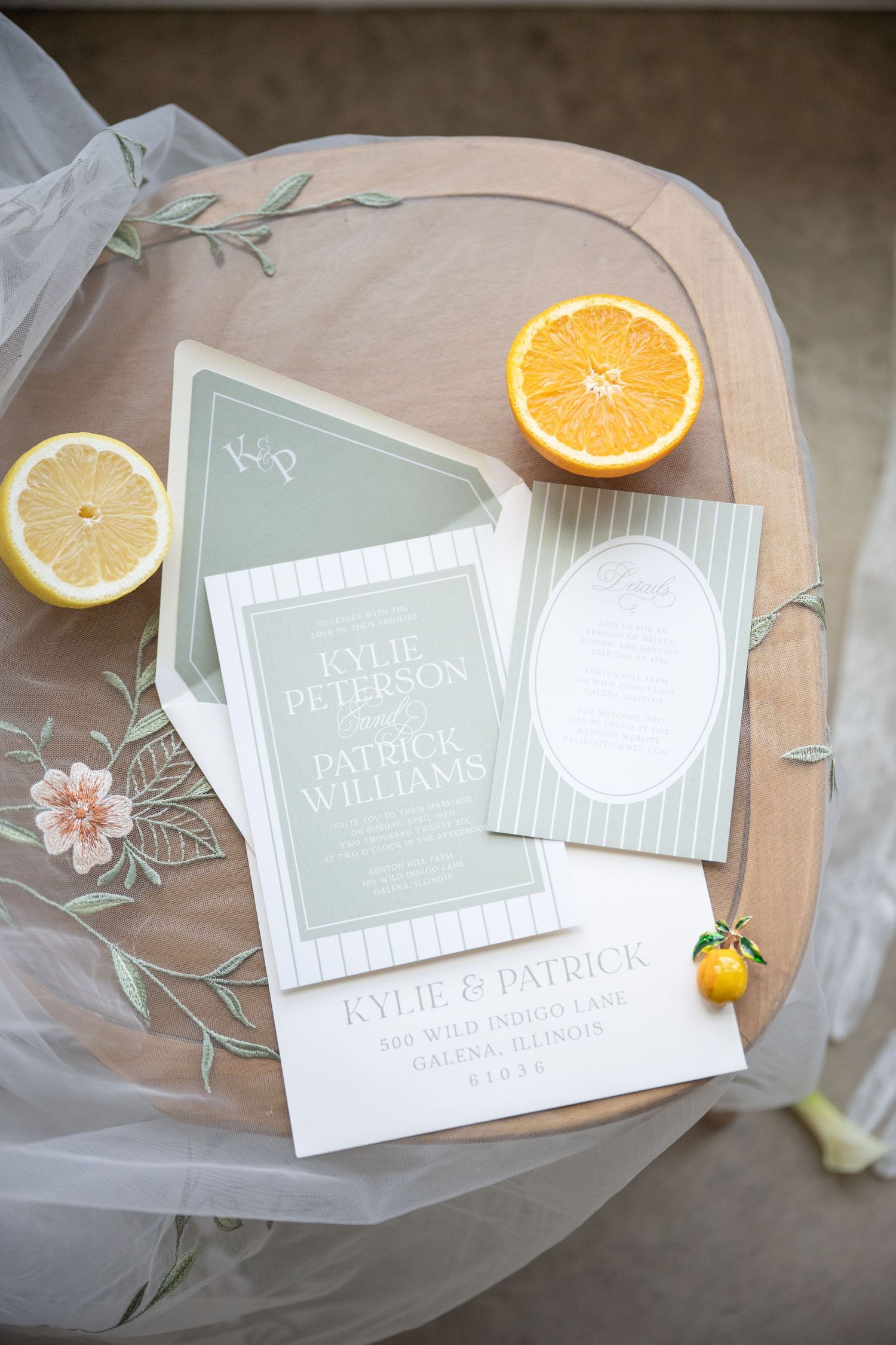 Sage green wedding invitation suite with striped details, displayed on a wooden table with fresh citrus fruit slices.