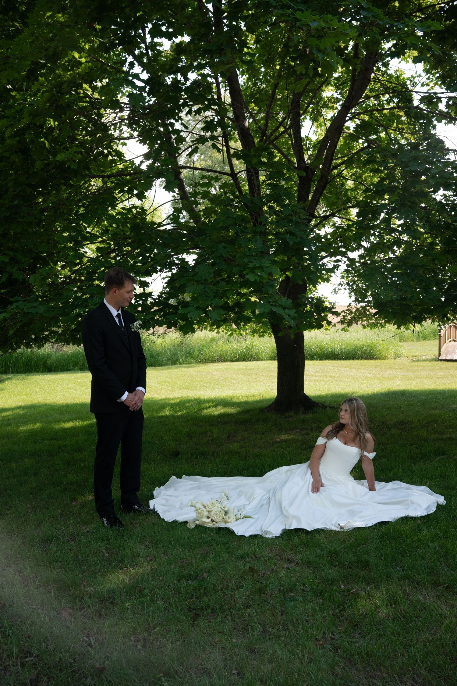 Groom in black tuxedo stands beside bride in white gown seated on grass under large trees.