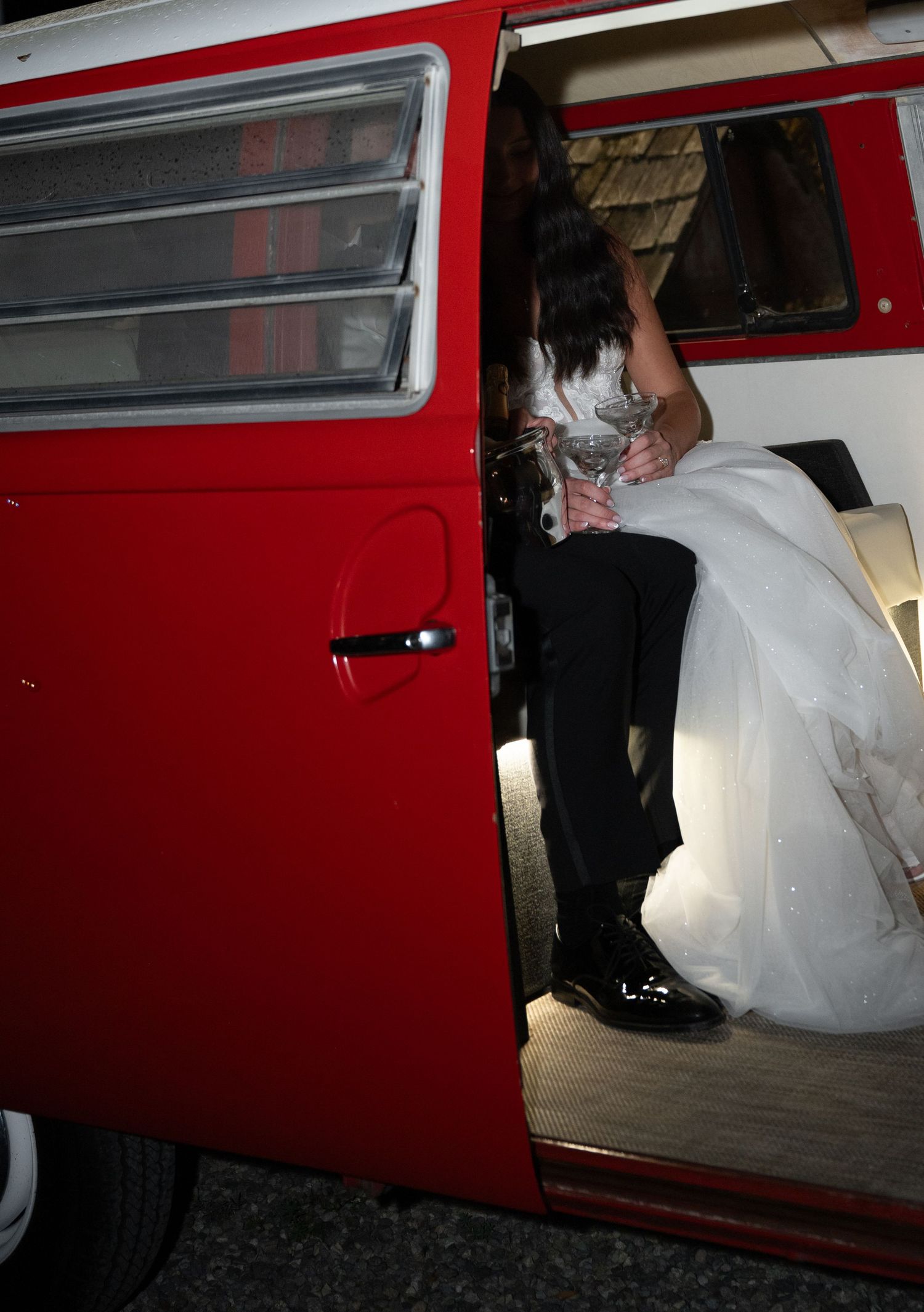Bride in white gown and groom share kiss inside red vintage Volkswagen wedding getaway van.