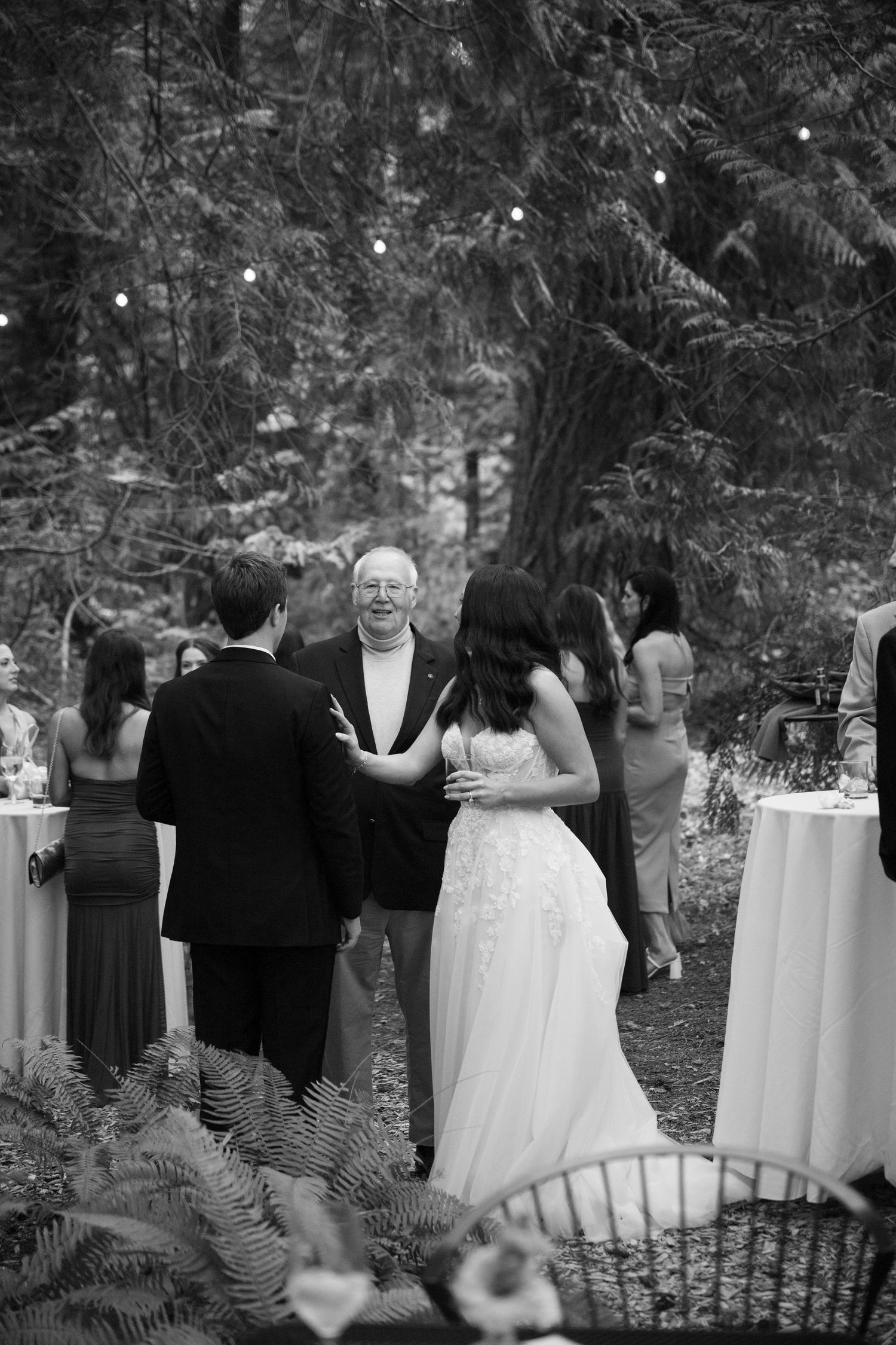 Black and white photo of bride and groom mingling with guests at outdoor wedding reception under string lights.