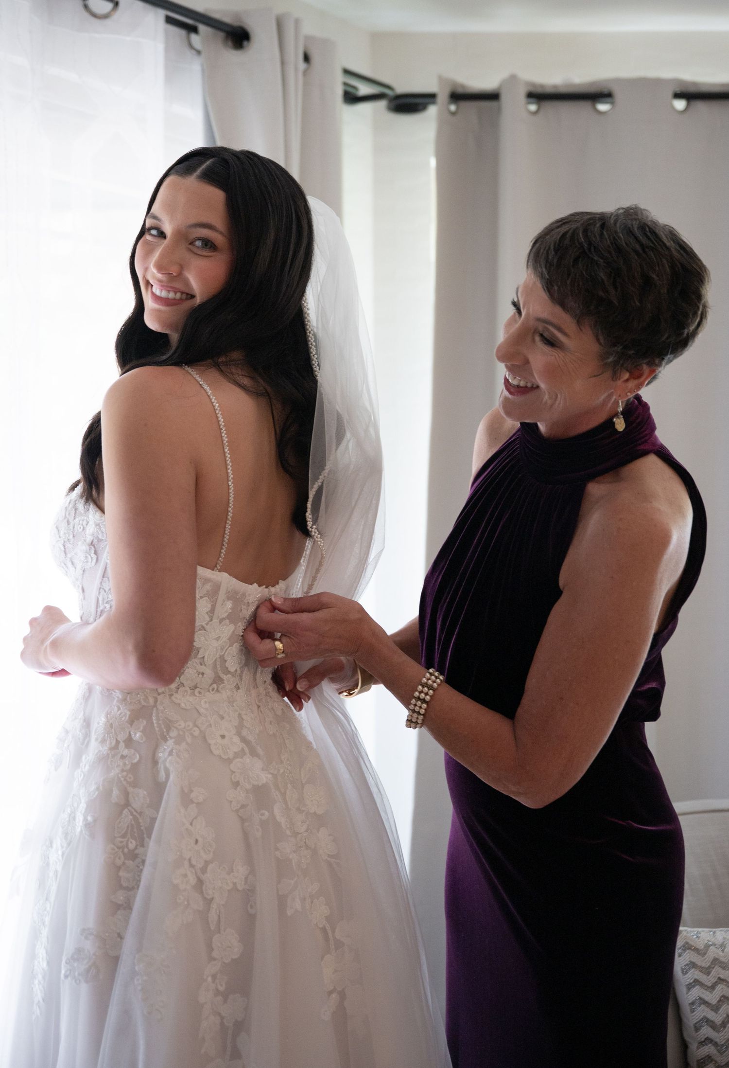 A bride in a white lace gown smiles as an older woman in purple helps fasten her wedding dress near a bright window.