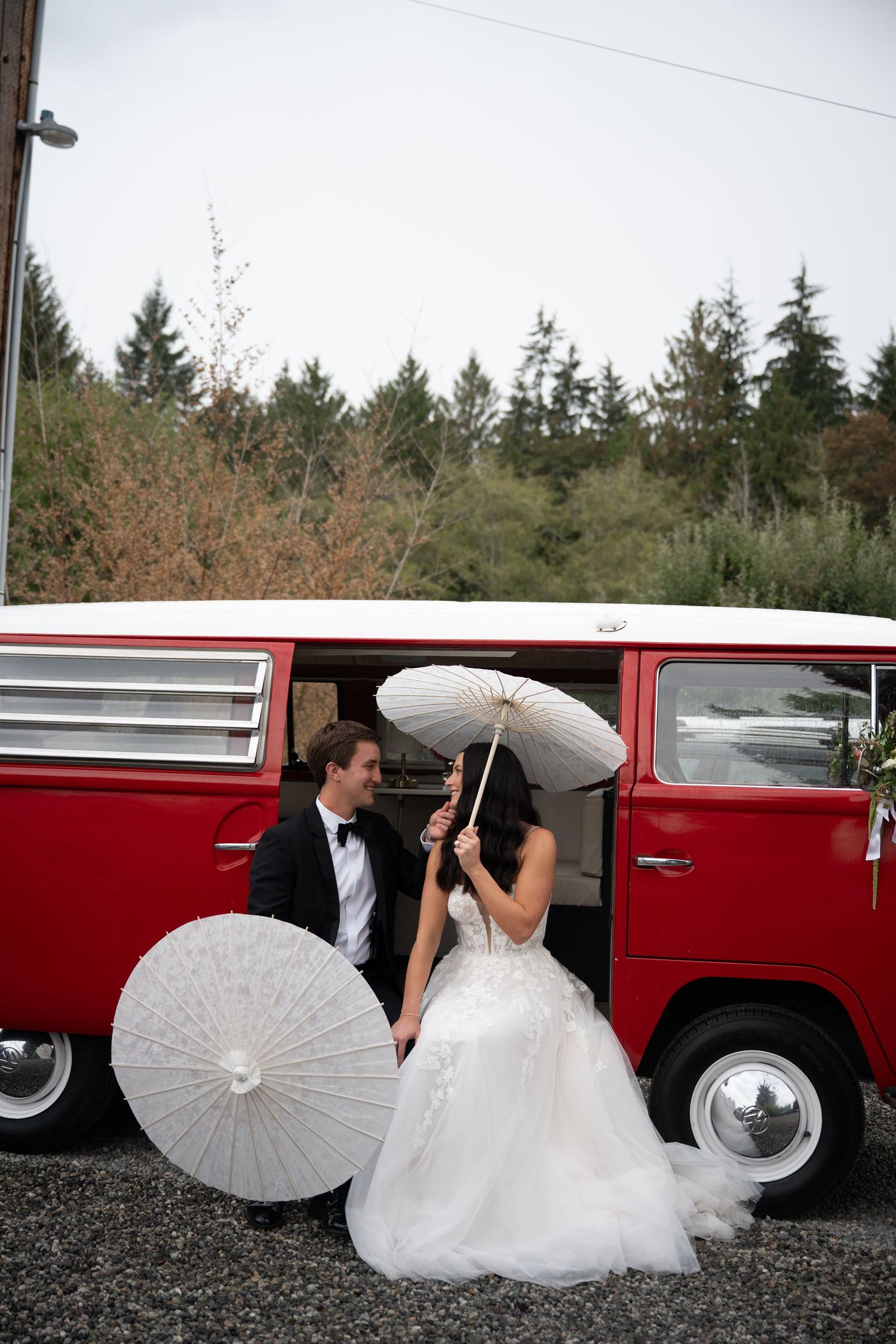 Bride and groom with white parasols beside a vintage red VW bus on their wedding day.