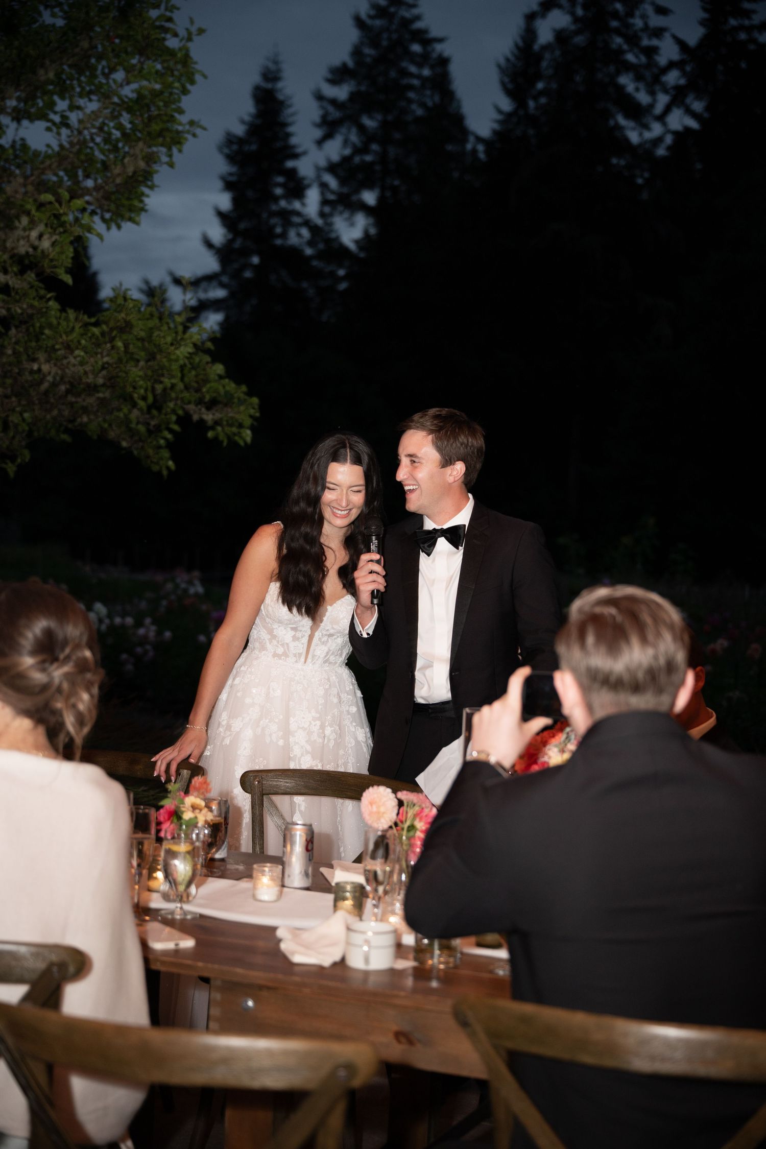 Bride and groom share a toast at an outdoor evening wedding reception surrounded by guests.
