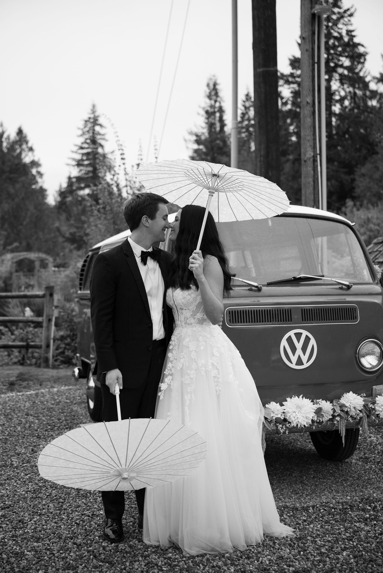 Black and white photo of newlyweds kissing with parasols in front of a classic VW van.