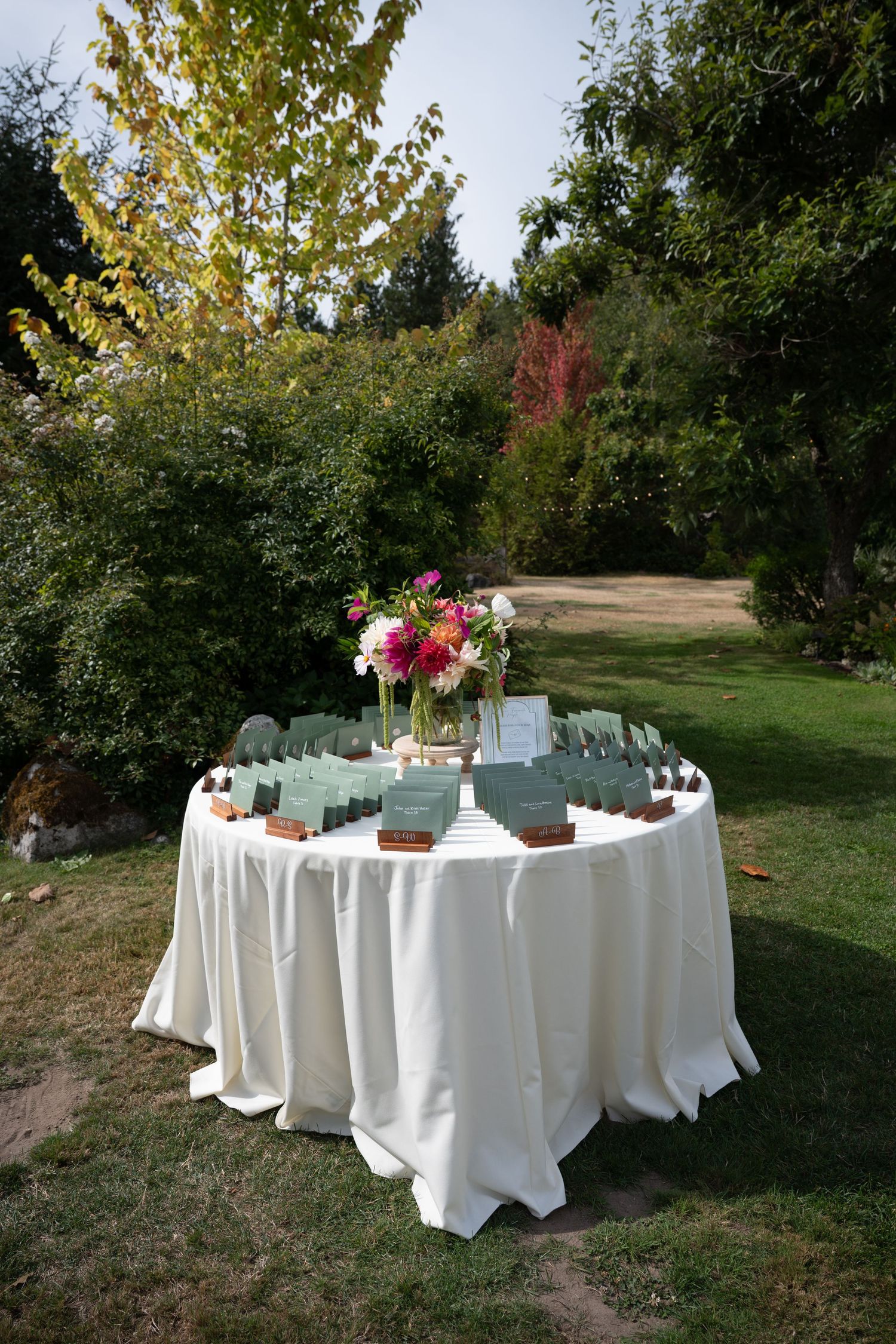 Outdoor wedding escort card table with white linen, green place cards, and pink floral centerpiece in a garden setting.