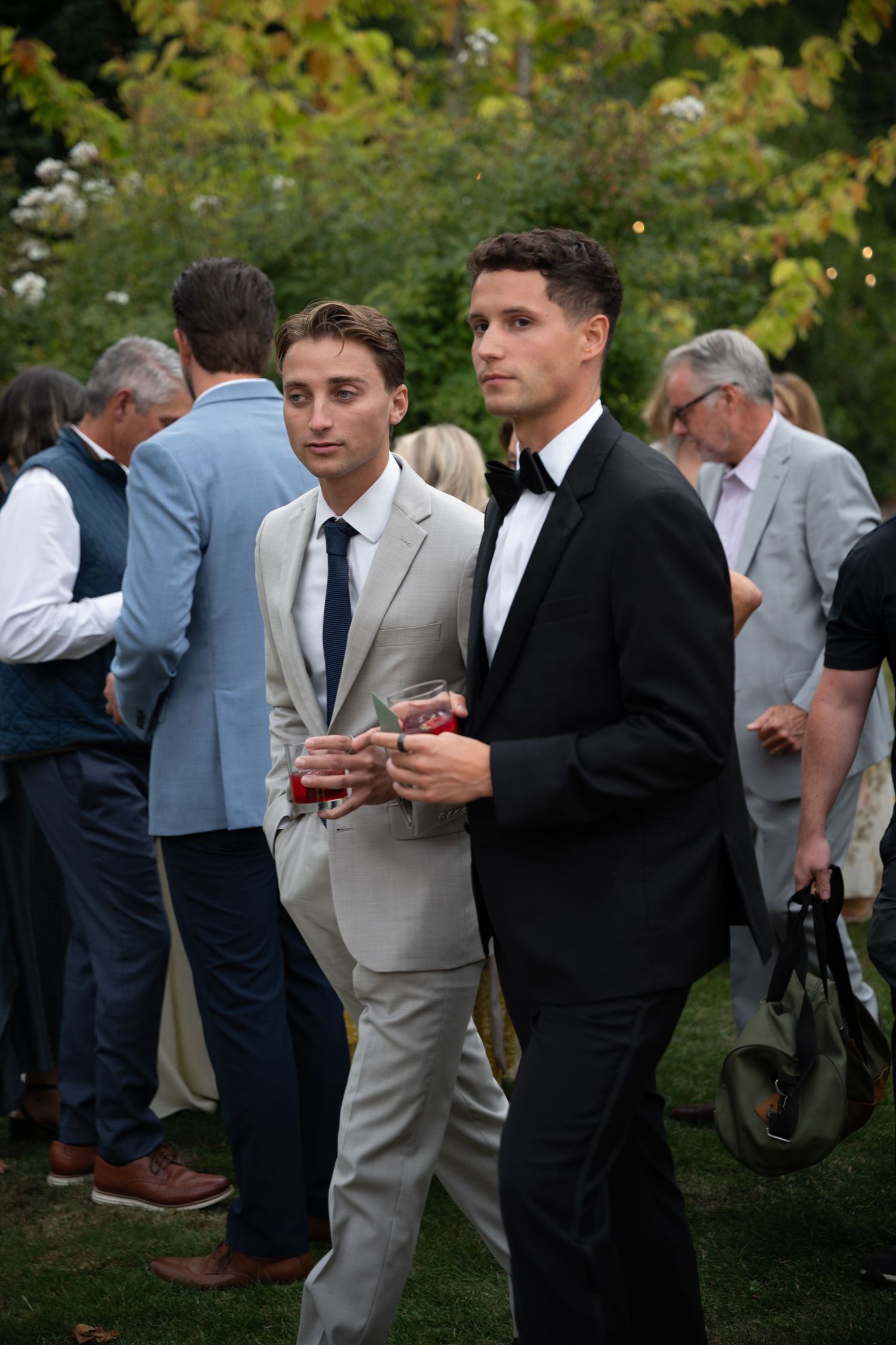 Two young men in suits, one beige one black tuxedo, stand at an outdoor wedding reception among guests.