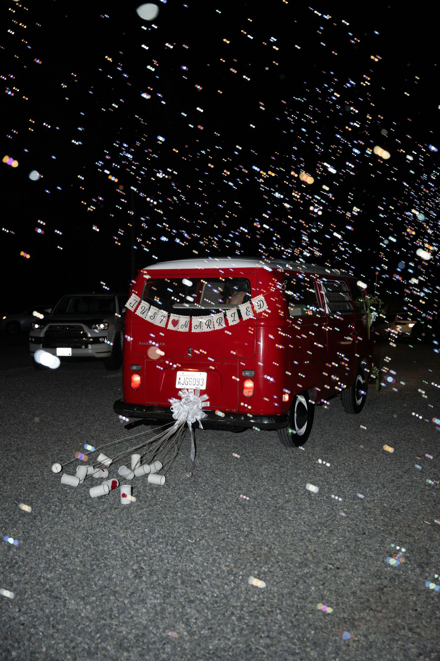 Red VW bus decorated with just married banner and tin cans, surrounded by confetti at night.
