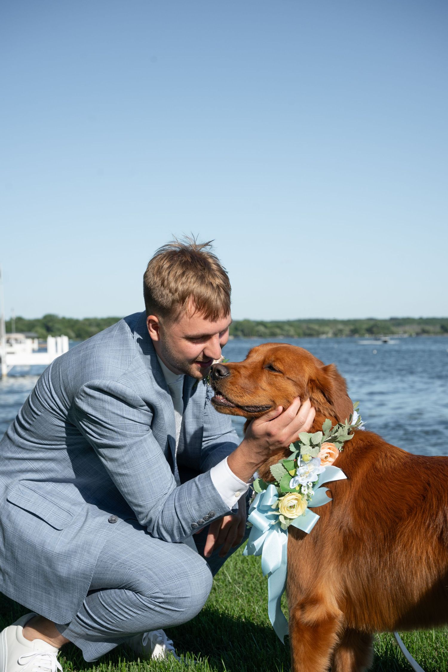 Groom in gray suit crouching to kiss his golden retriever dog wearing a floral collar at waterfront wedding.