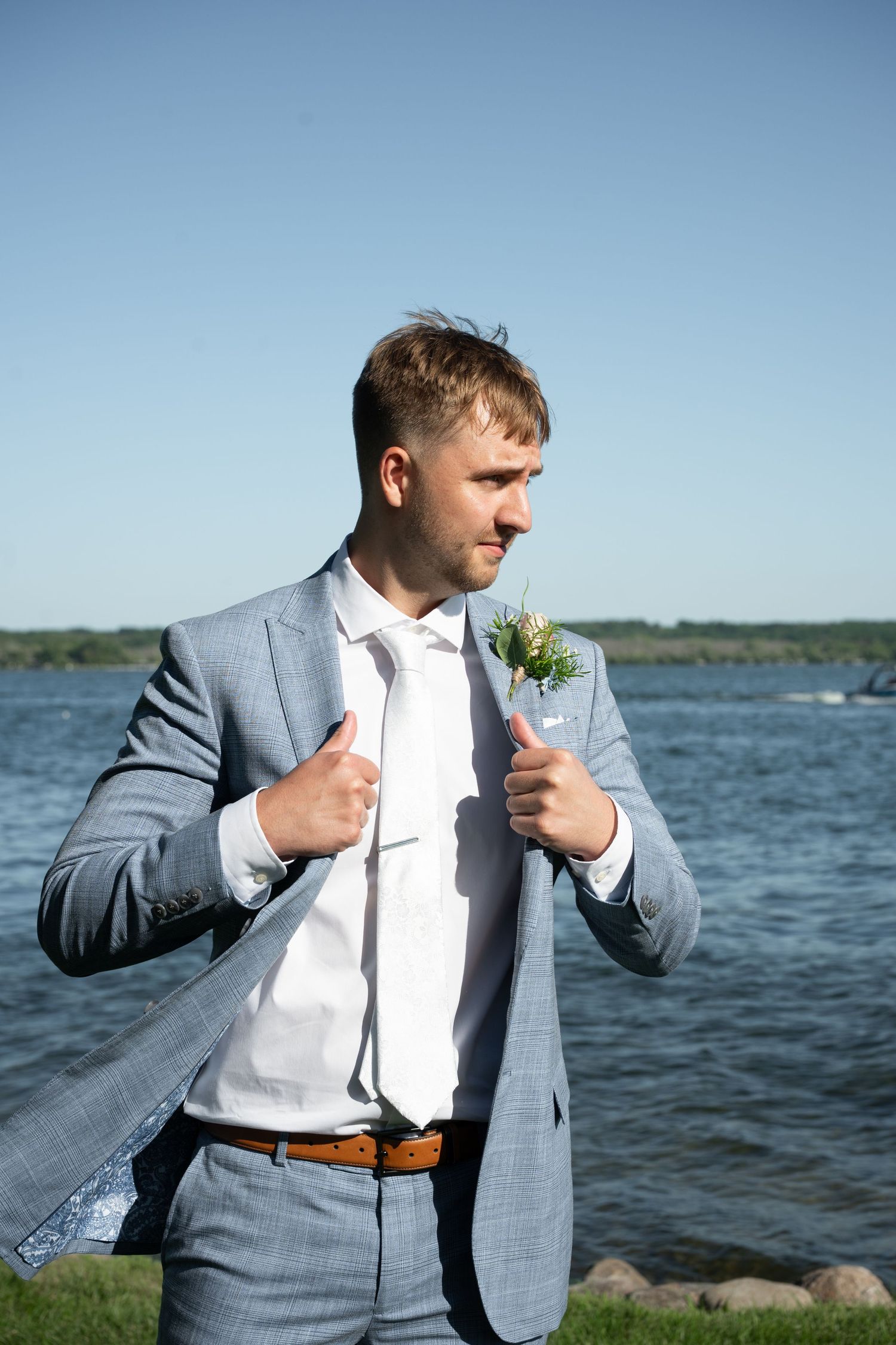 Groom in gray suit with boutonniere poses confidently by waterfront on sunny wedding day.