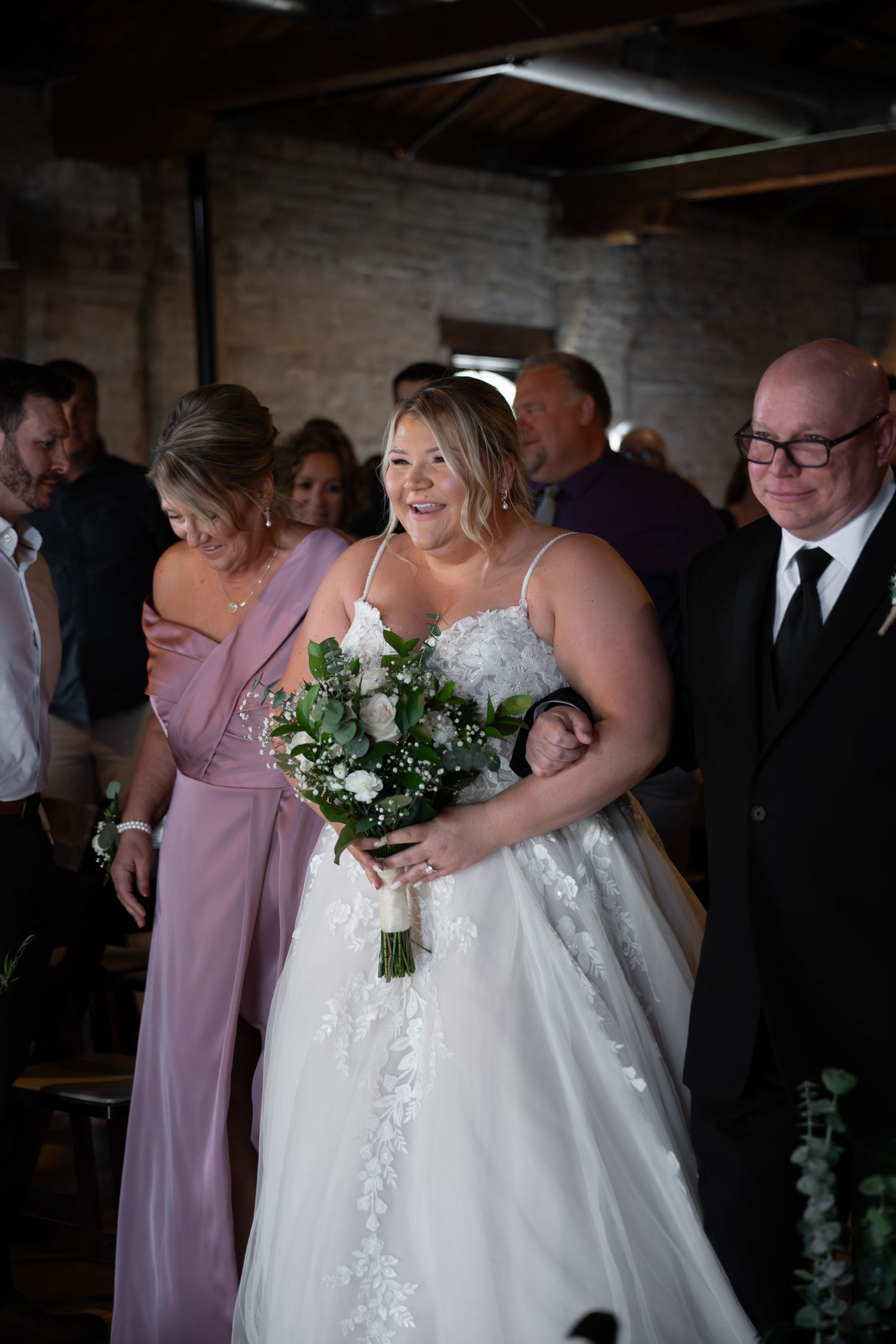 Smiling bride in lace wedding gown walks down aisle holding bouquet, escorted by family members.