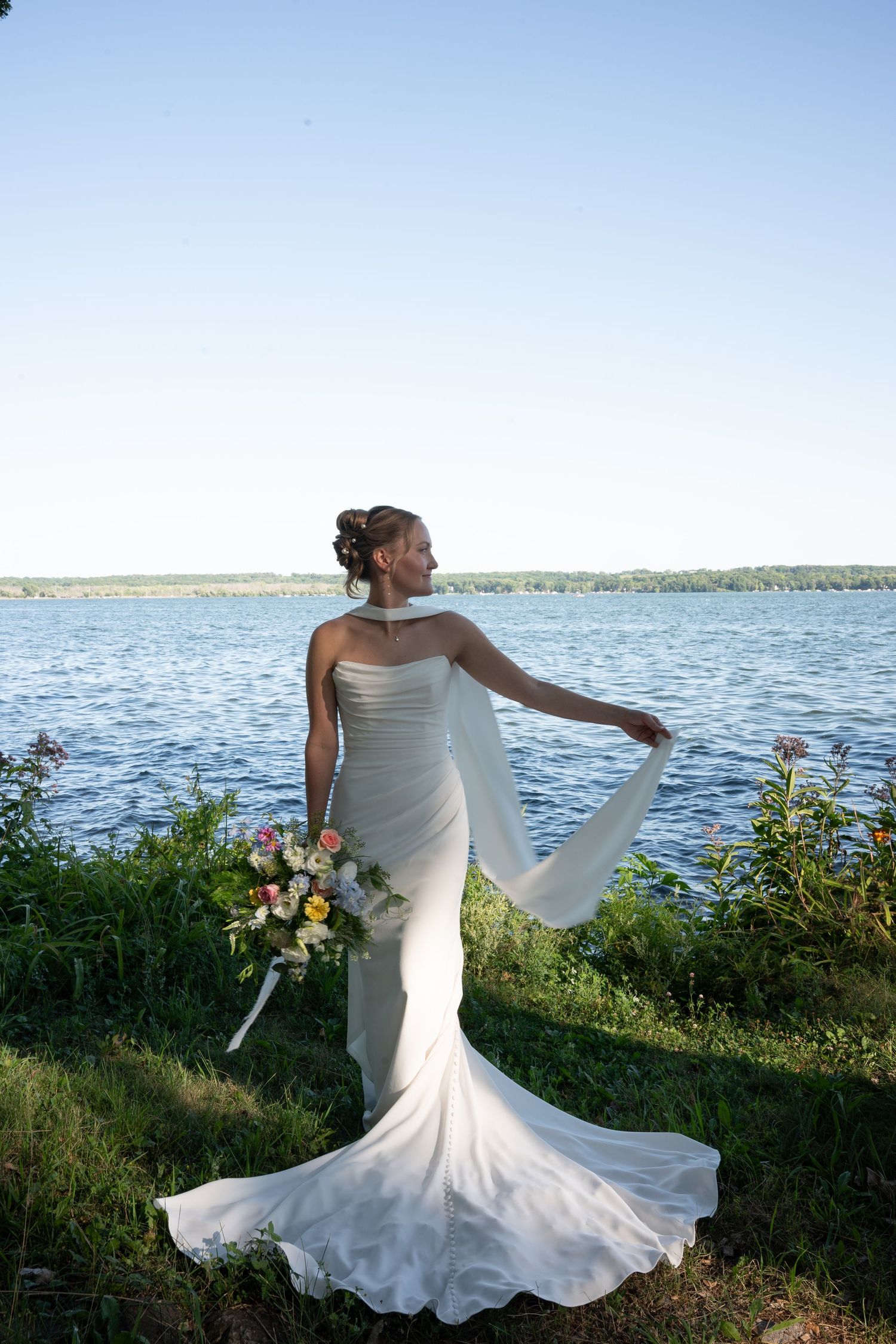 Bride in white mermaid gown holding bouquet and flowing veil by a serene lakeside on a sunny day.