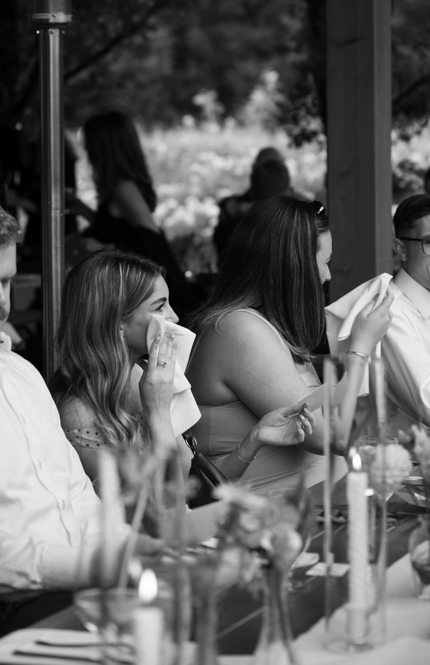 Black and white photo of emotional wedding guests wiping tears at an outdoor reception dinner table.