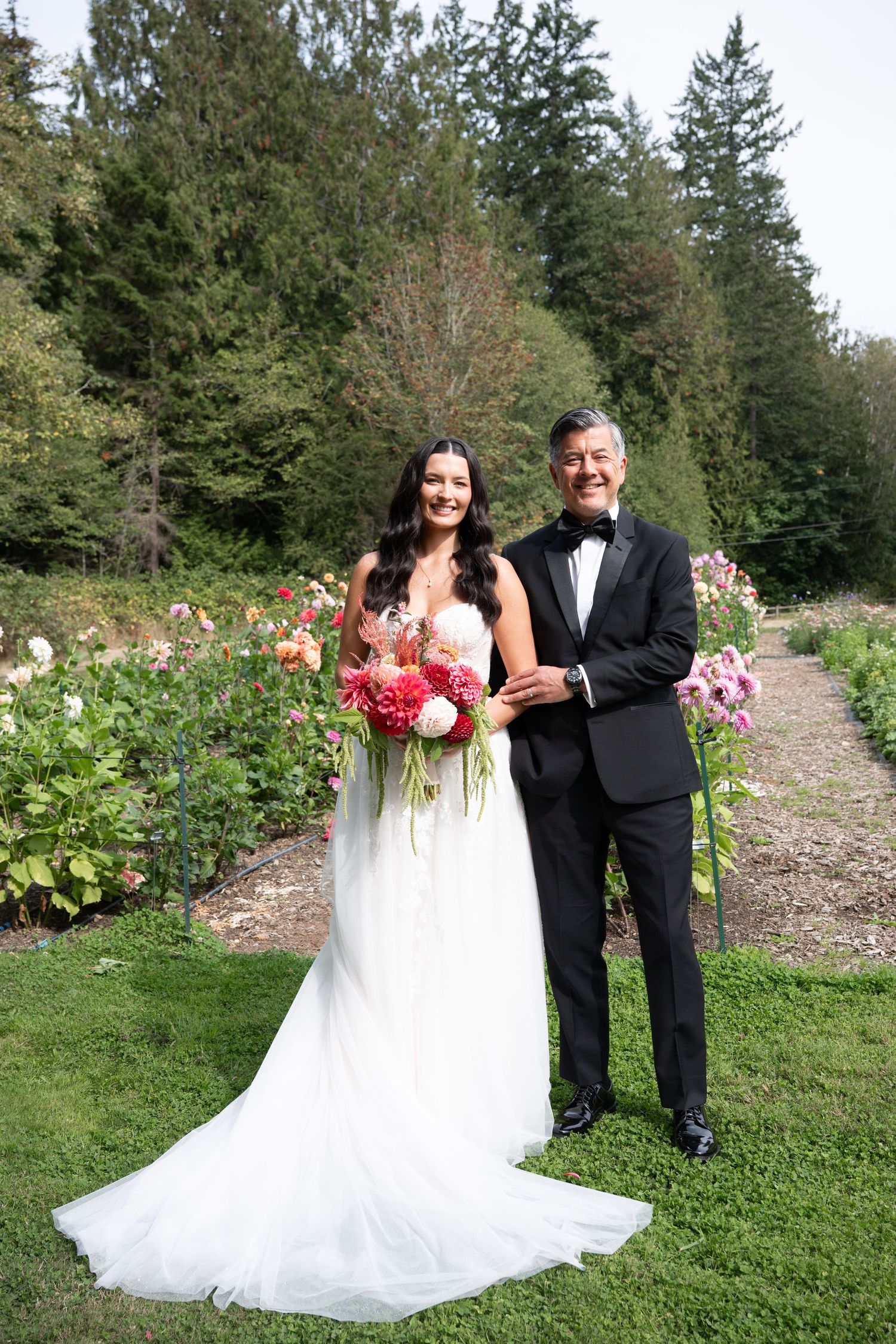 Bride and groom pose together in a garden wedding setting, surrounded by colorful dahlias, bride holding pink bouquet.