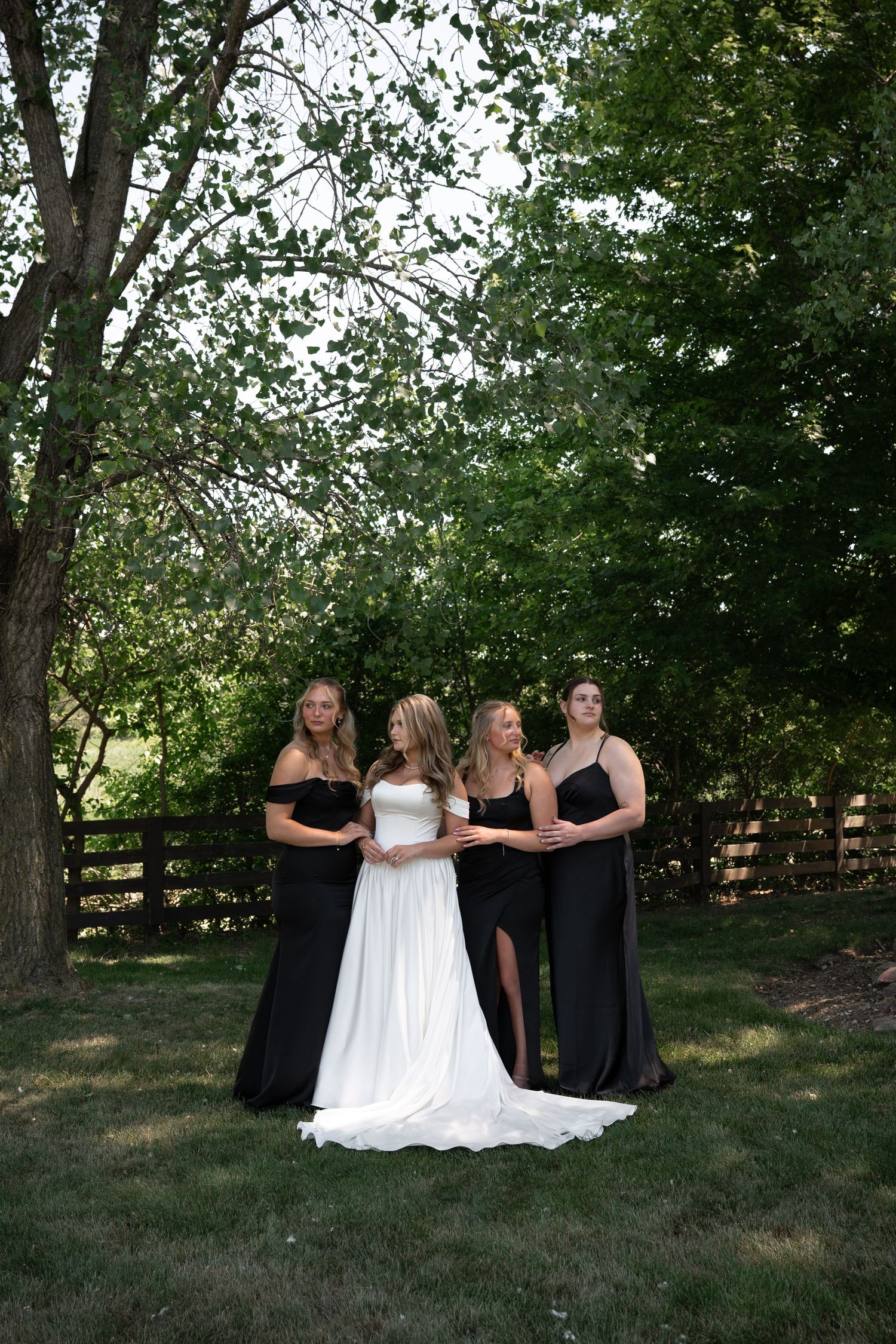 Bride in white gown poses with three bridesmaids in black dresses under green trees outdoors.