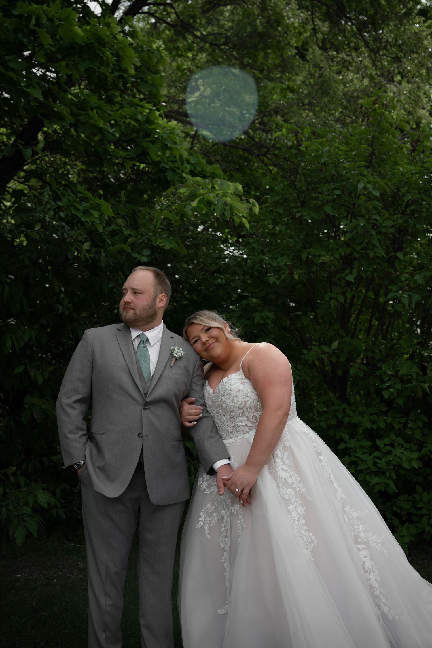 Bride in white ballgown leans on groom in gray suit surrounded by vibrant green trees.