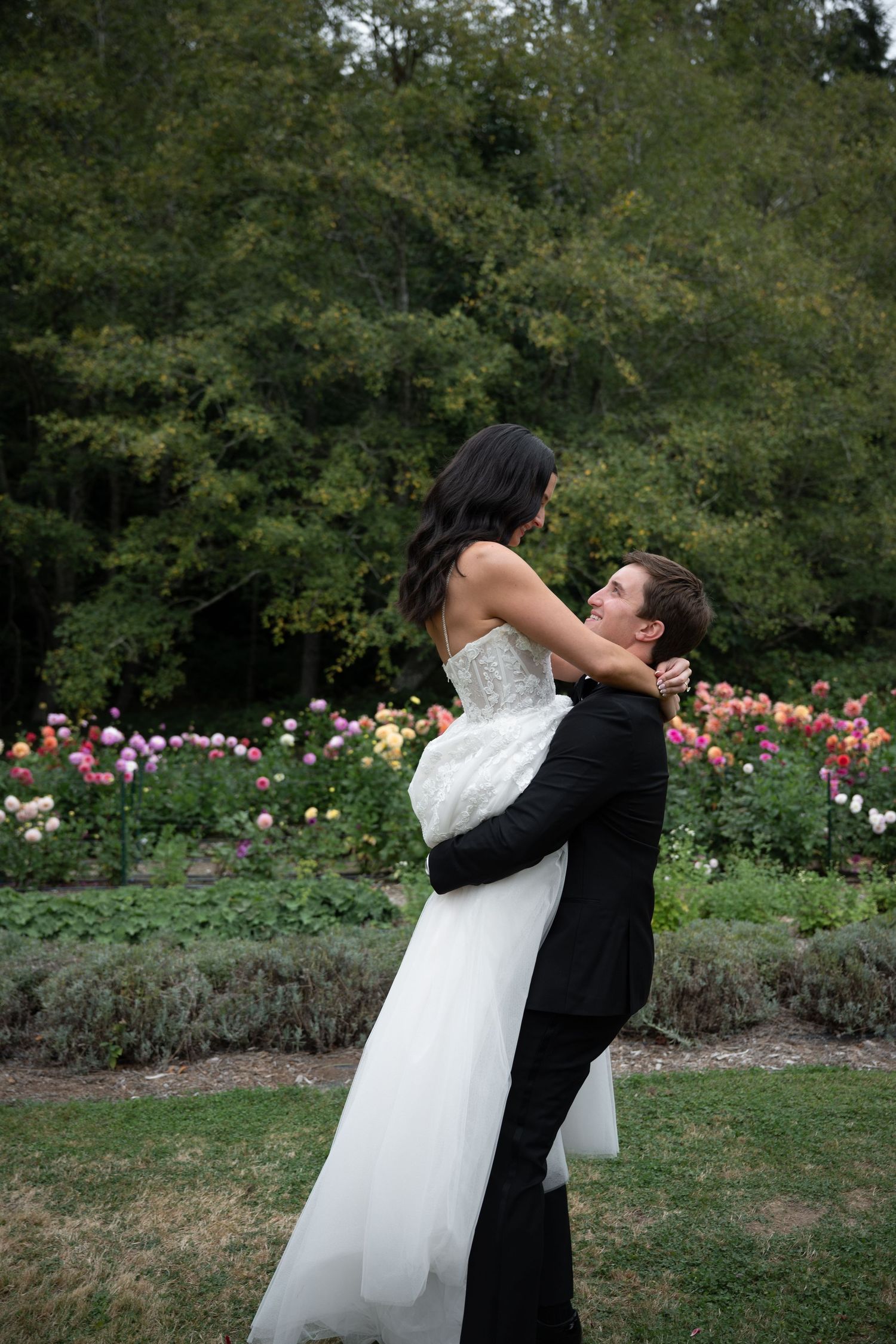 Groom lifts bride in white strapless gown while embracing in front of colorful flower garden.