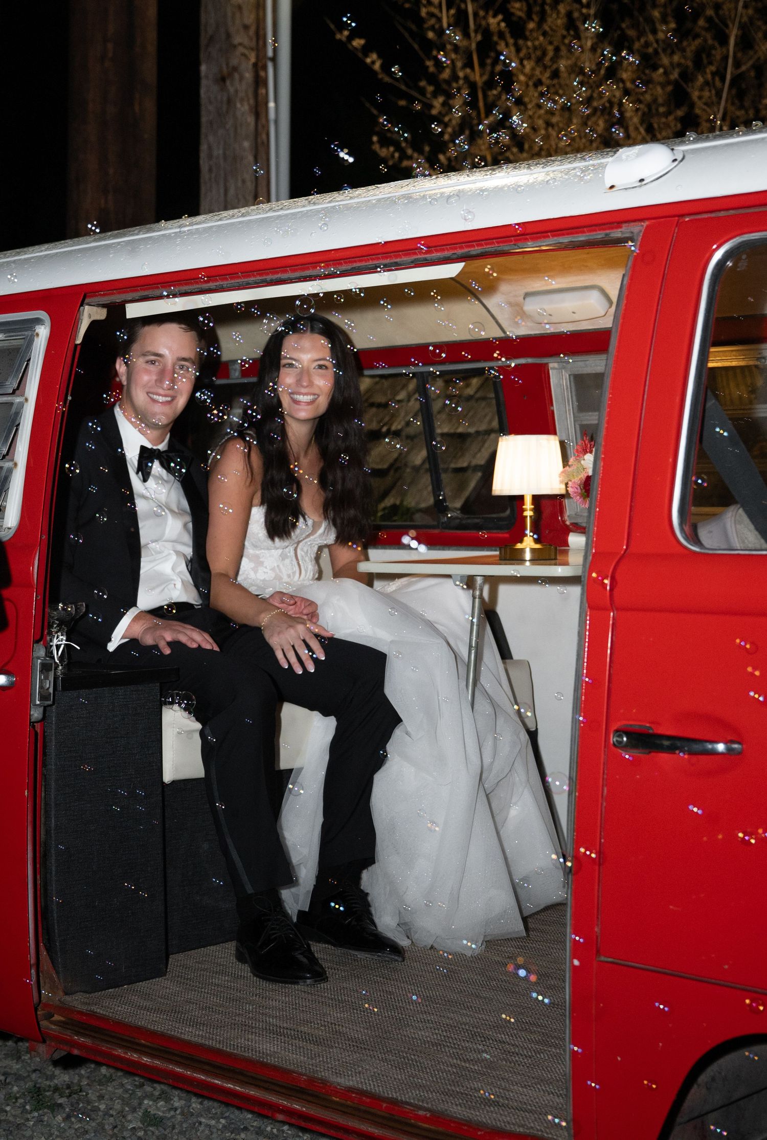 Smiling bride and groom sit inside decorated vintage red Volkswagen van at night wedding.