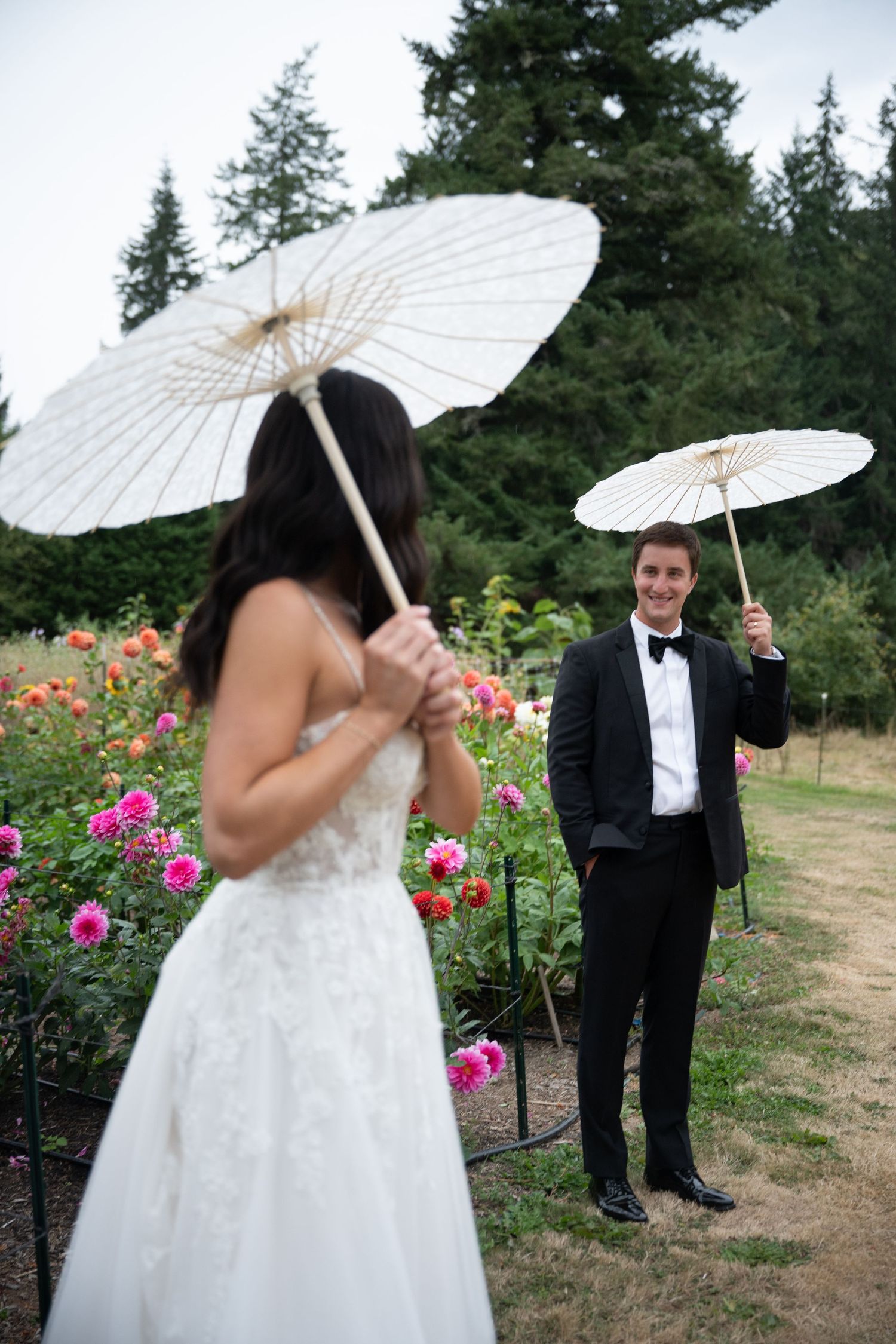 Bride and groom hold white parasols in a colorful garden during their outdoor wedding ceremony.