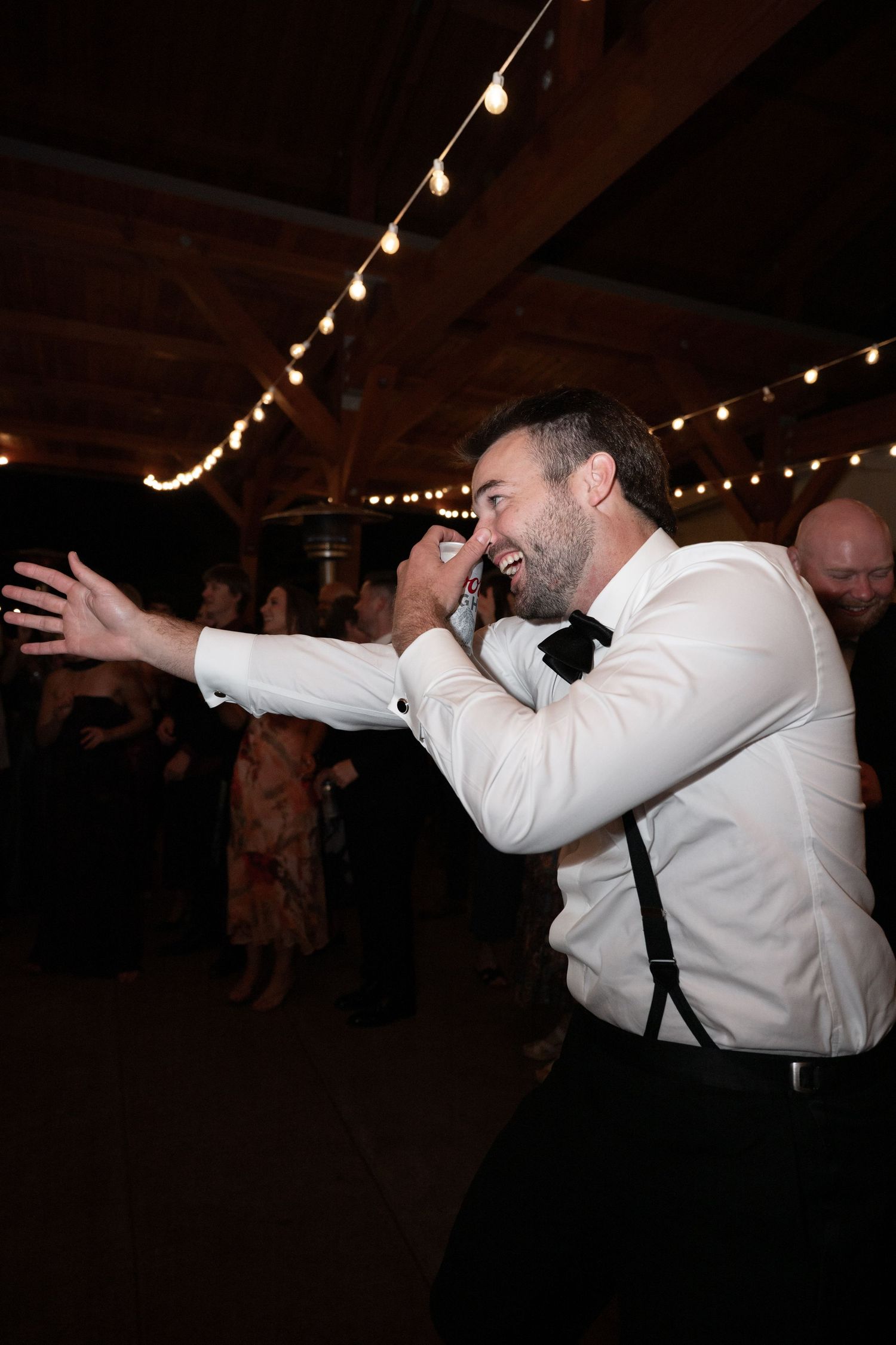 Energetic man in white shirt and suspenders dancing at wedding reception with string lights.