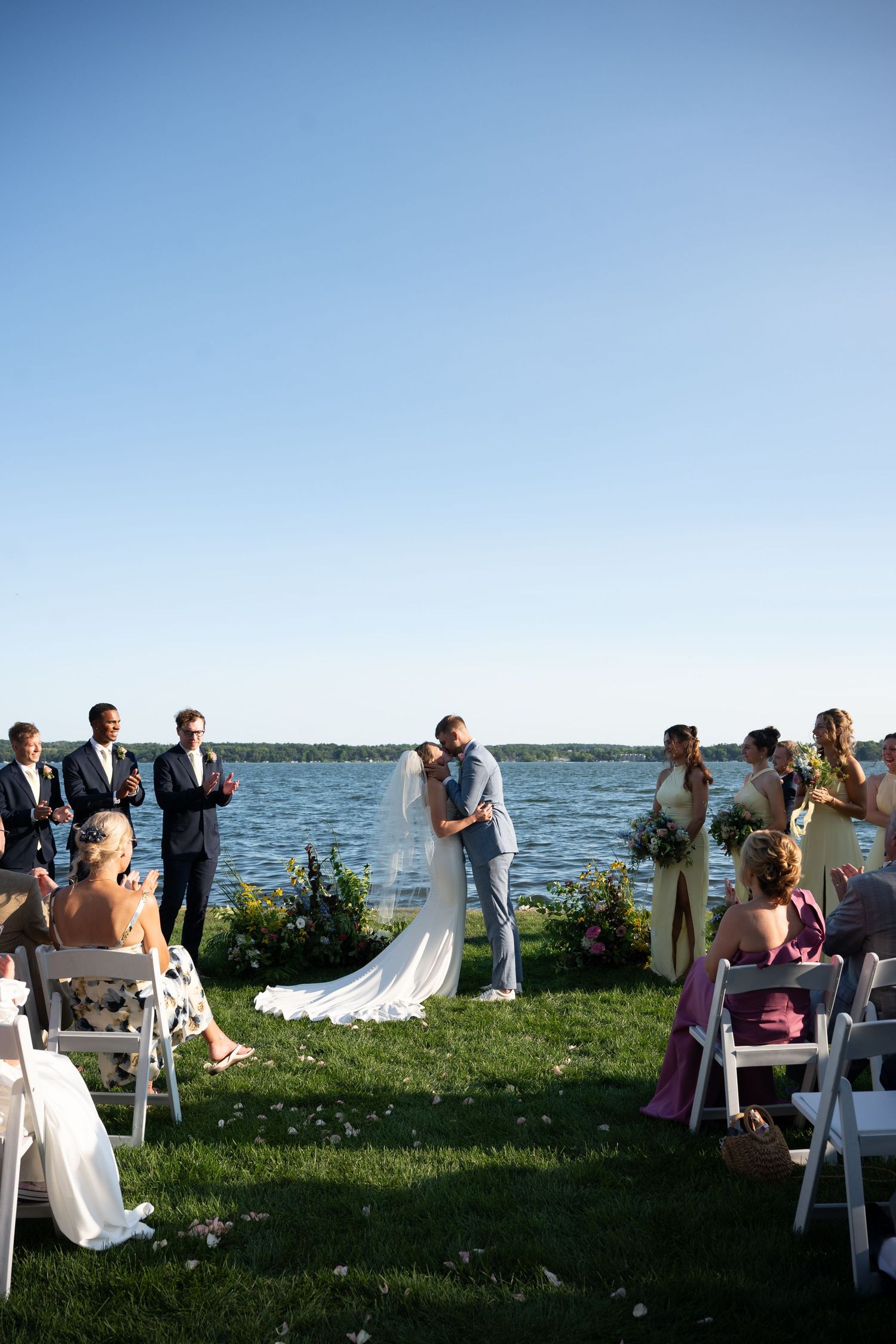 Bride and groom share first kiss at outdoor lakeside wedding ceremony with guests seated on white chairs.