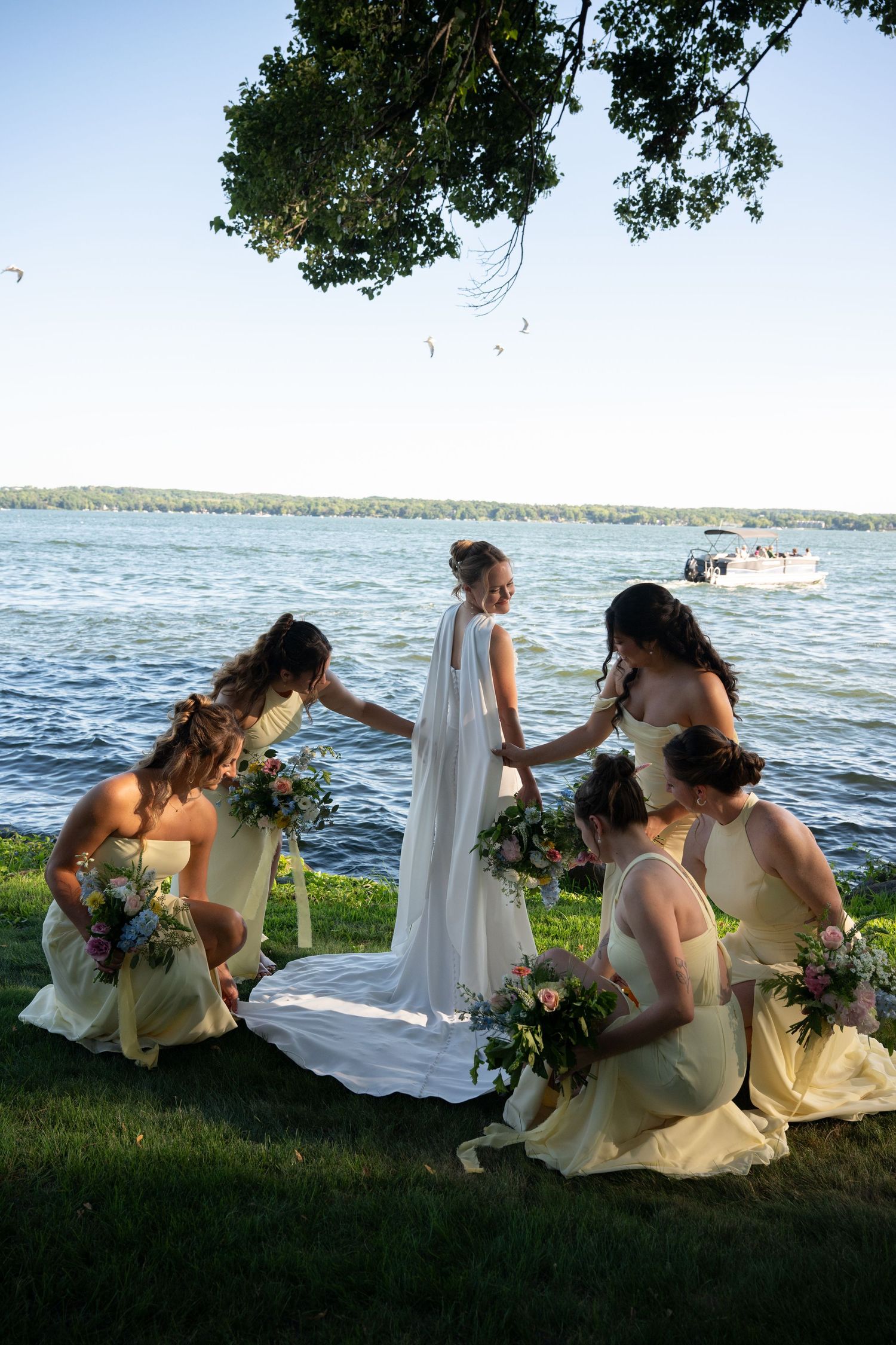 Bridesmaids in yellow dresses kneel around a bride in a white gown by a serene lakeside on her wedding day.