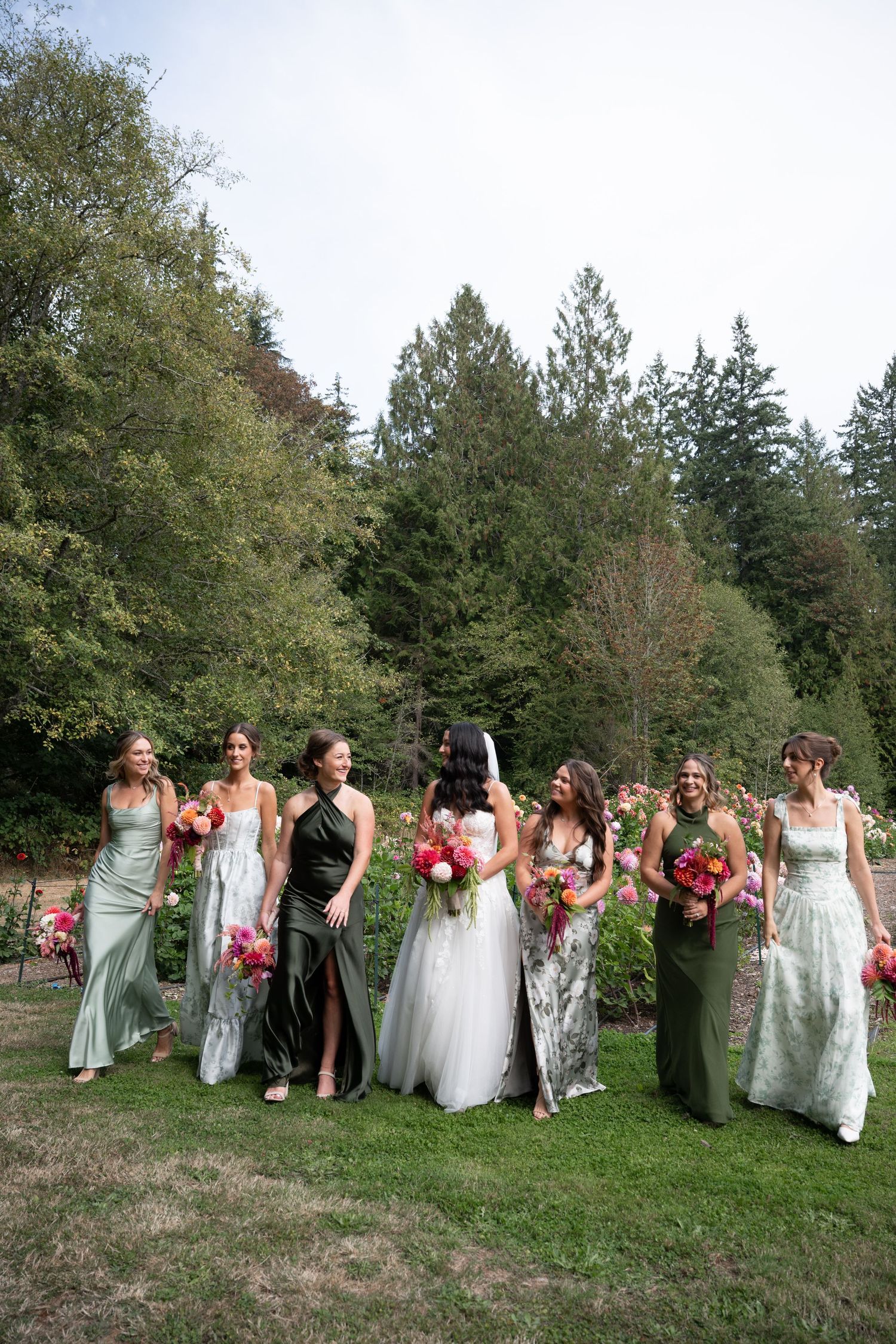 Bride and bridesmaids in mixed green gowns walk together holding colorful bouquets in a lush garden setting.