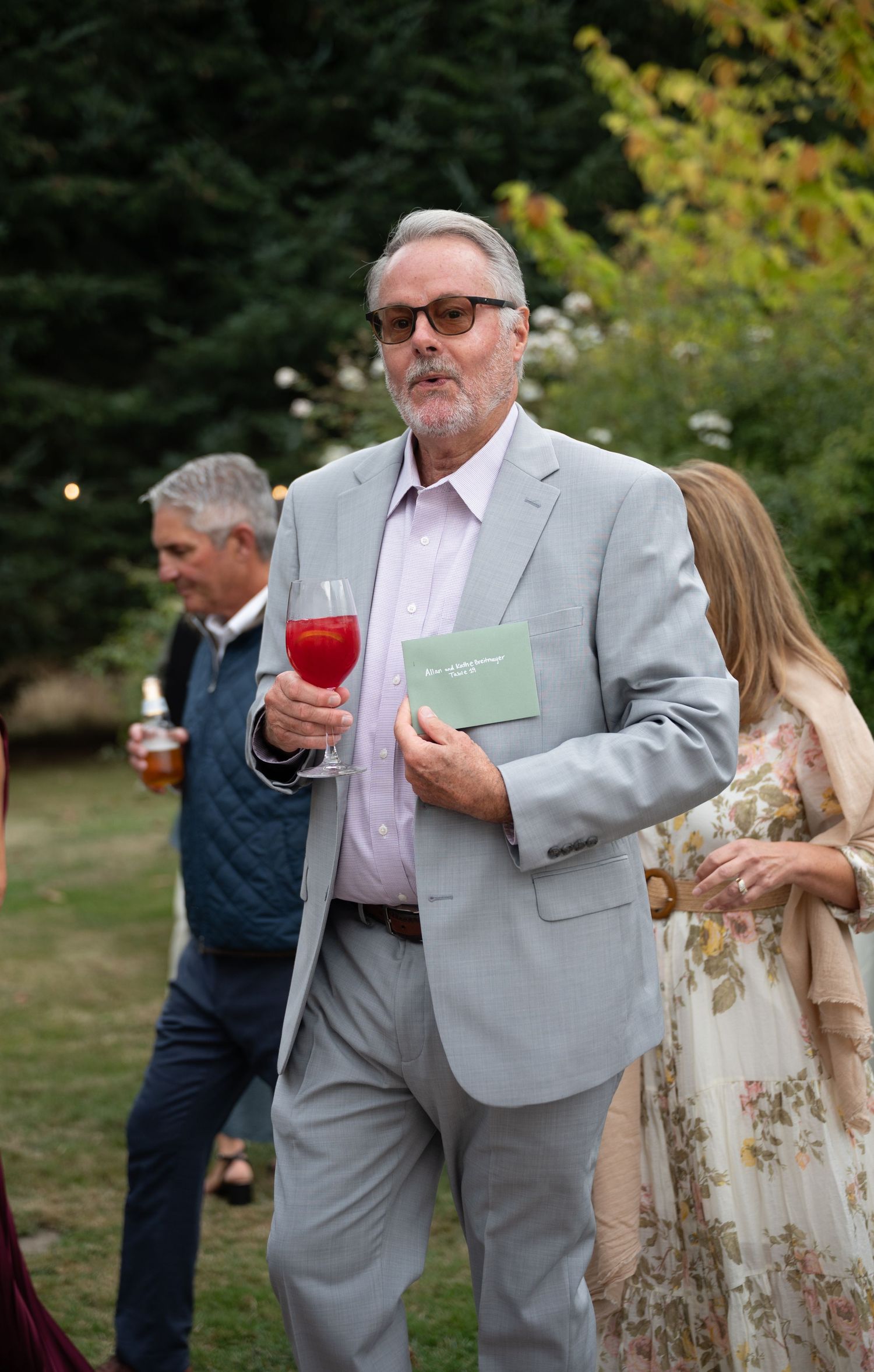 Wedding guest in light gray suit holding red drink and green program booklet at outdoor garden reception.