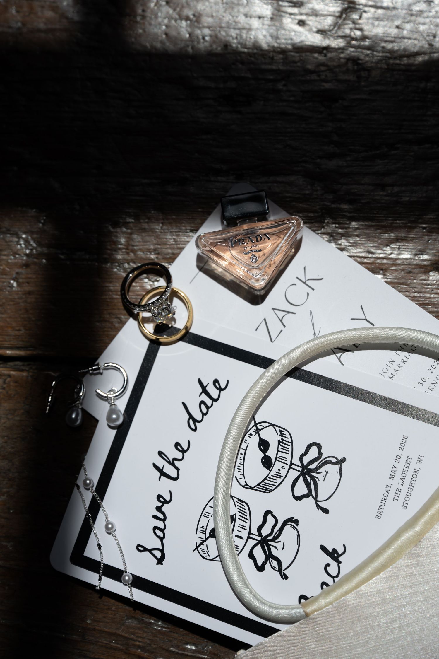 Save the date wedding card with gold rings, pearl bracelet, and Prada perfume on rustic wood.
